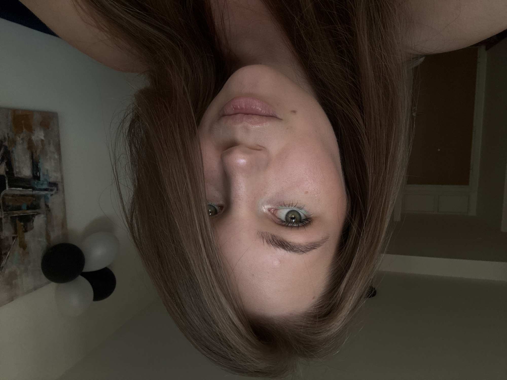 A medium shot captures a young woman from the shoulders up, facing the camera. She has long, light brown hair that frames her face. Her eyes are green, and she is wearing light makeup, including a touch of teal eyeliner. A small mole is visible on her left cheek. The background is dimly lit, with a sliver of a white wall to her right, adorned with a framed abstract painting and a cluster of black and white balloons. To her left, a dark doorway is visible. The overall lighting is soft and diffused. The scene appears to be indoors, possibly in a residential setting. There is no specific indicator of the city of Belgrade, Serbia.