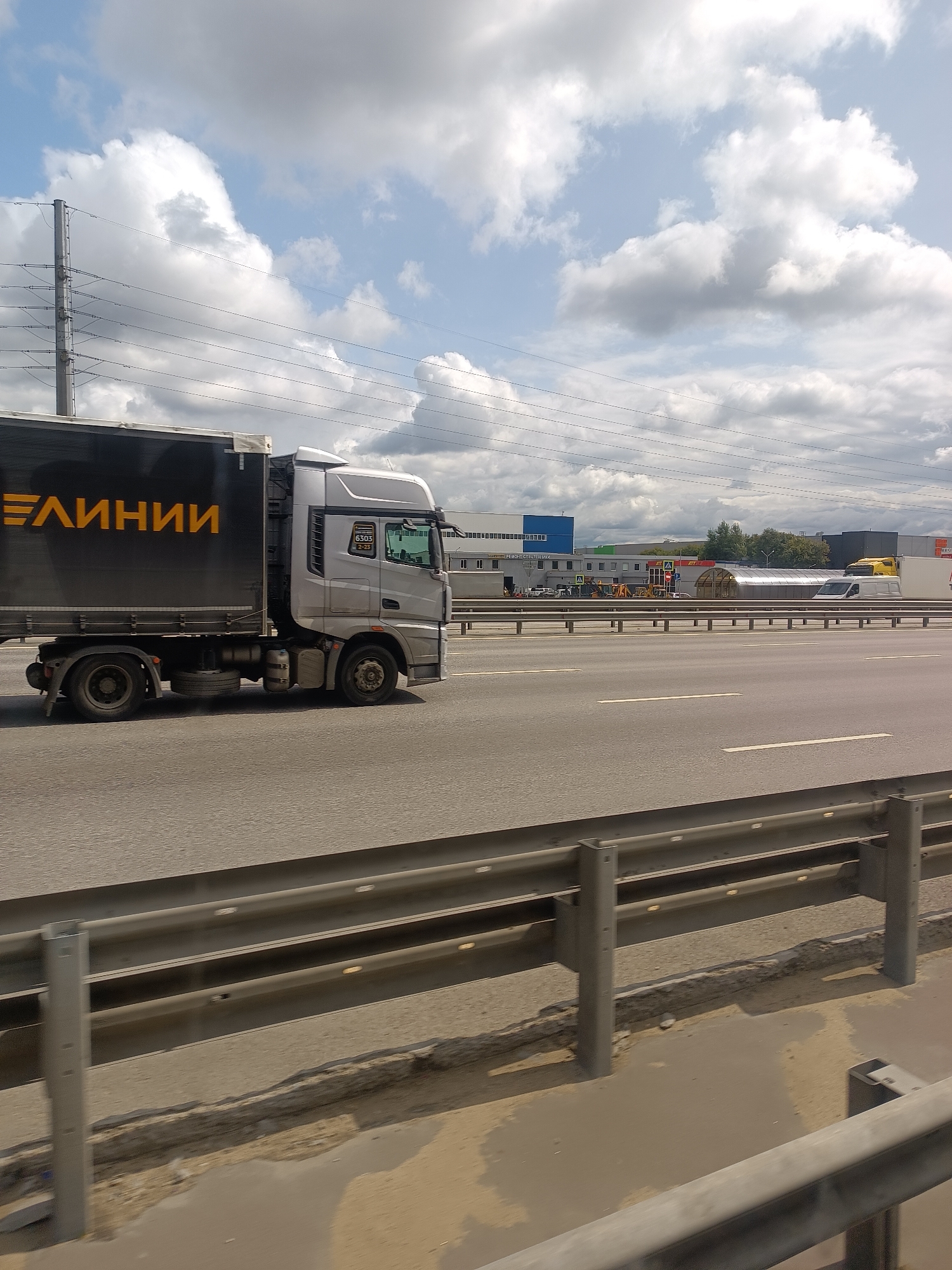 A silver semi-trailer truck is traveling on a multi-lane highway. The truck has a black trailer with yellow lettering that reads "ЕЛЕНИНИИ". The truck's cab has the number "6303" and "2-23" displayed on a small sign. The highway has a metal guardrail on the right side of the image. In the background, there are several industrial buildings and a tall structure with power lines. The sky is partly cloudy. The image was taken in Tomilino, Russia.