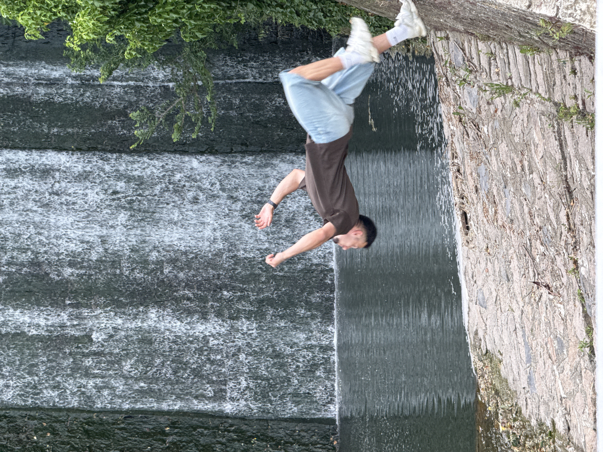 A man is doing a flip in front of a waterfall. The man is wearing a brown t-shirt, blue shorts, white socks, and white shoes. The waterfall is tiered, with green foliage growing at the top. A stone wall is visible to the right of the waterfall. The image is rotated 90 degrees clockwise. This image was taken at Waterfall in Sea Garden, Varna, Bulgaria.