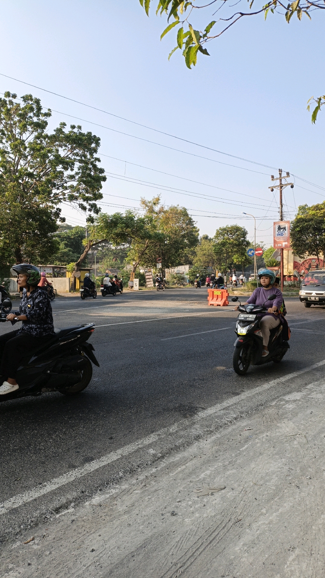 The image depicts a street scene in Sumbring, Indonesia, likely a suburban or semi-urban environment. The time of day appears to be late afternoon, with warm sunlight casting long shadows. The sky is clear and blue, with a few power lines crisscrossing overhead.
The foreground is dominated by a paved road with white lane markings. On the left side of the road, a woman wearing a helmet and a patterned shirt is riding a black scooter. She is looking forward, and her posture suggests she is in motion. To her right, on the same road, another woman wearing a purple shirt and a light blue helmet is riding a black scooter with a passenger on the back. They are also looking forward, suggesting they are moving through the intersection.
In the background, there are several other people on scooters, indicating active traffic. Trees with lush green foliage line the sides of the road, and some buildings are visible behind them. Orange traffic barriers are placed on the road, suggesting some form of roadwork or traffic management. A signpost with an IND logo and directional information is also visible. A black car is parked or moving on the right side of the road. The road surface shows signs of wear and tear, with some patches of loose gravel and tire marks. The overall impression is a typical day in a moderately busy Indonesian street.