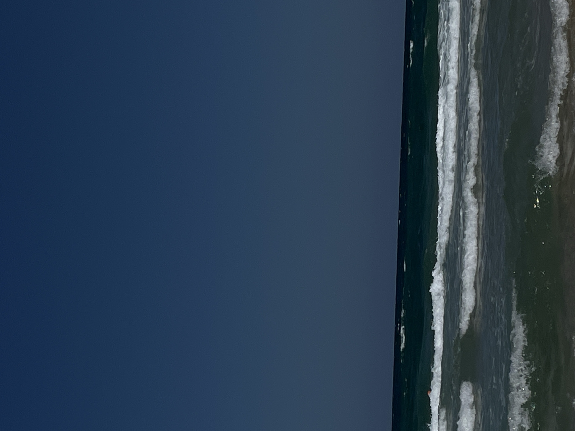 The image captures a wide, serene expanse of the sea under a deep blue sky. The horizon line, where the dark blue of the sky meets the dark green of the sea, is sharply defined. The sea is characterized by several rows of white-capped waves rolling towards the viewer. The water transitions from a deep, dark green in the distance to a more translucent, lighter green closer to the foreground, with hints of sand visible at the very bottom edge of the frame. A small, orange object, possibly a buoy or a piece of debris, is visible in the water to the left of the frame, amidst the darker green water. There are no discernible people or any structures in the scene. The overall impression is one of natural tranquility and the vastness of the ocean. This scene could be representative of the coastline of Primorsko, Bulgaria, known for its beaches and clear sea waters.