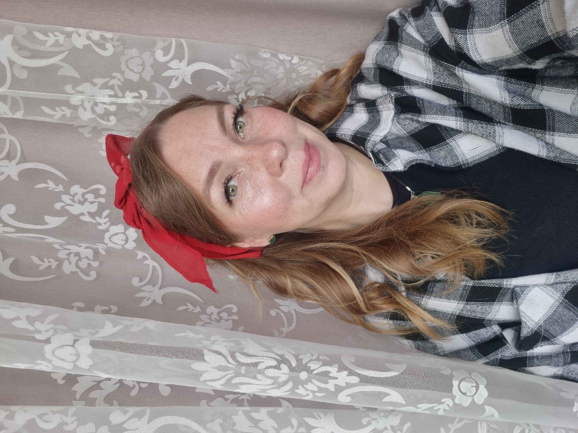A woman with fair skin, freckles, and green eyes is lying on a patterned white surface. She has reddish-brown hair, with a red ribbon tied in it. She is wearing a black top and a black and white checkered flannel shirt. The photo appears to have been taken indoors. The location of Chaikovsky, Russia cannot be confirmed without more information or evidence in the image.