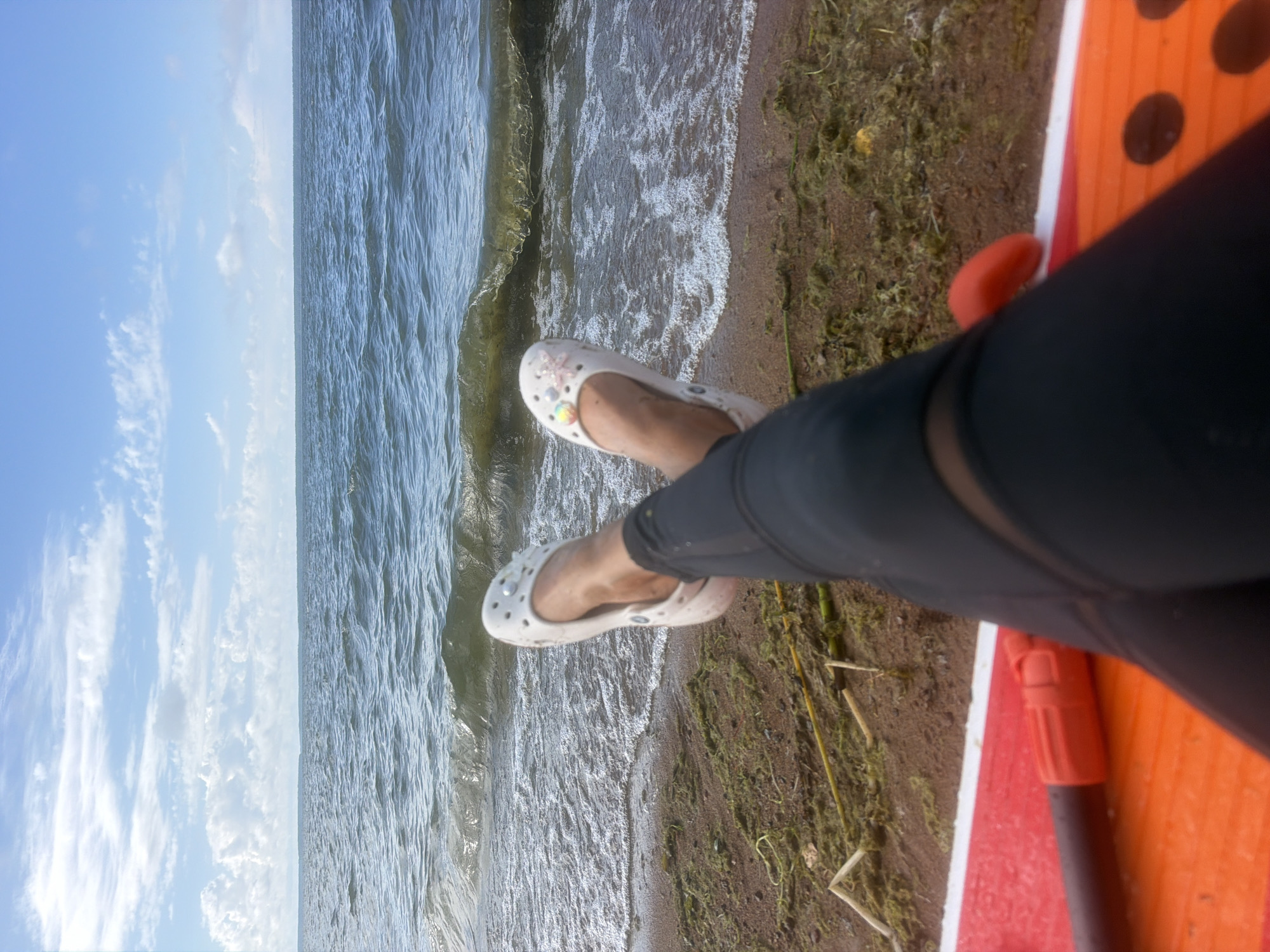 The image depicts a person's legs and feet, adorned with white Crocs, resting on the edge of an orange paddleboard. The person is wearing black leggings with a mesh panel detail. They are positioned on a sandy shore, where green algae and reeds are visible. The scene overlooks a body of water with gentle waves lapping at the shore. The sky above is a clear blue, dotted with white clouds. The location is the city of Vyazy, Russia.