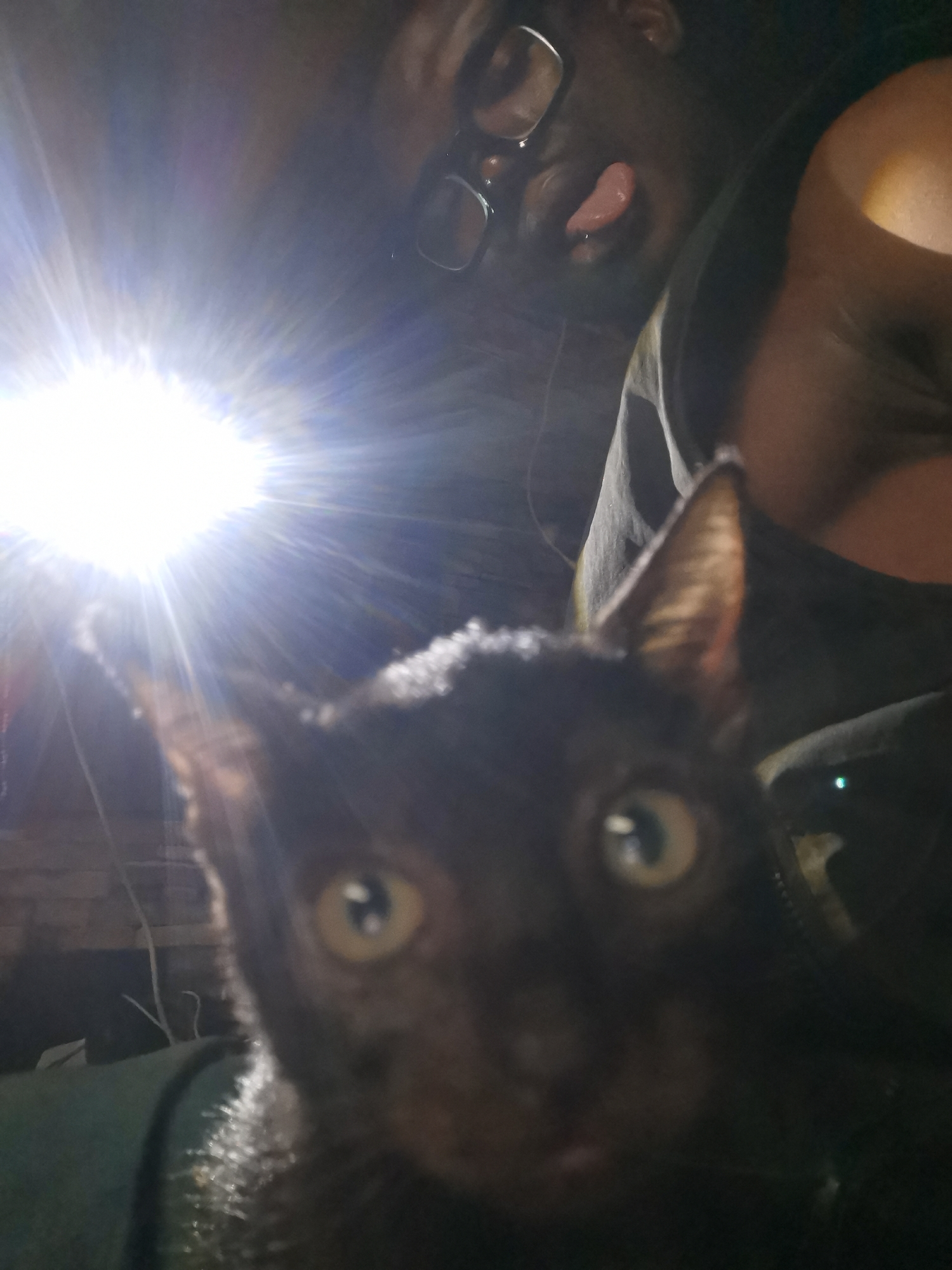 The image shows a dark-skinned person wearing glasses sticking their tongue out. In front of the person is a dark-colored cat with large, round eyes. A bright light source is positioned to the left of the frame, obscuring details of the background. Clothing appears to be a dark-colored sleeveless garment. No elements are discernable to indicate the location is Lagos, Nigeria.