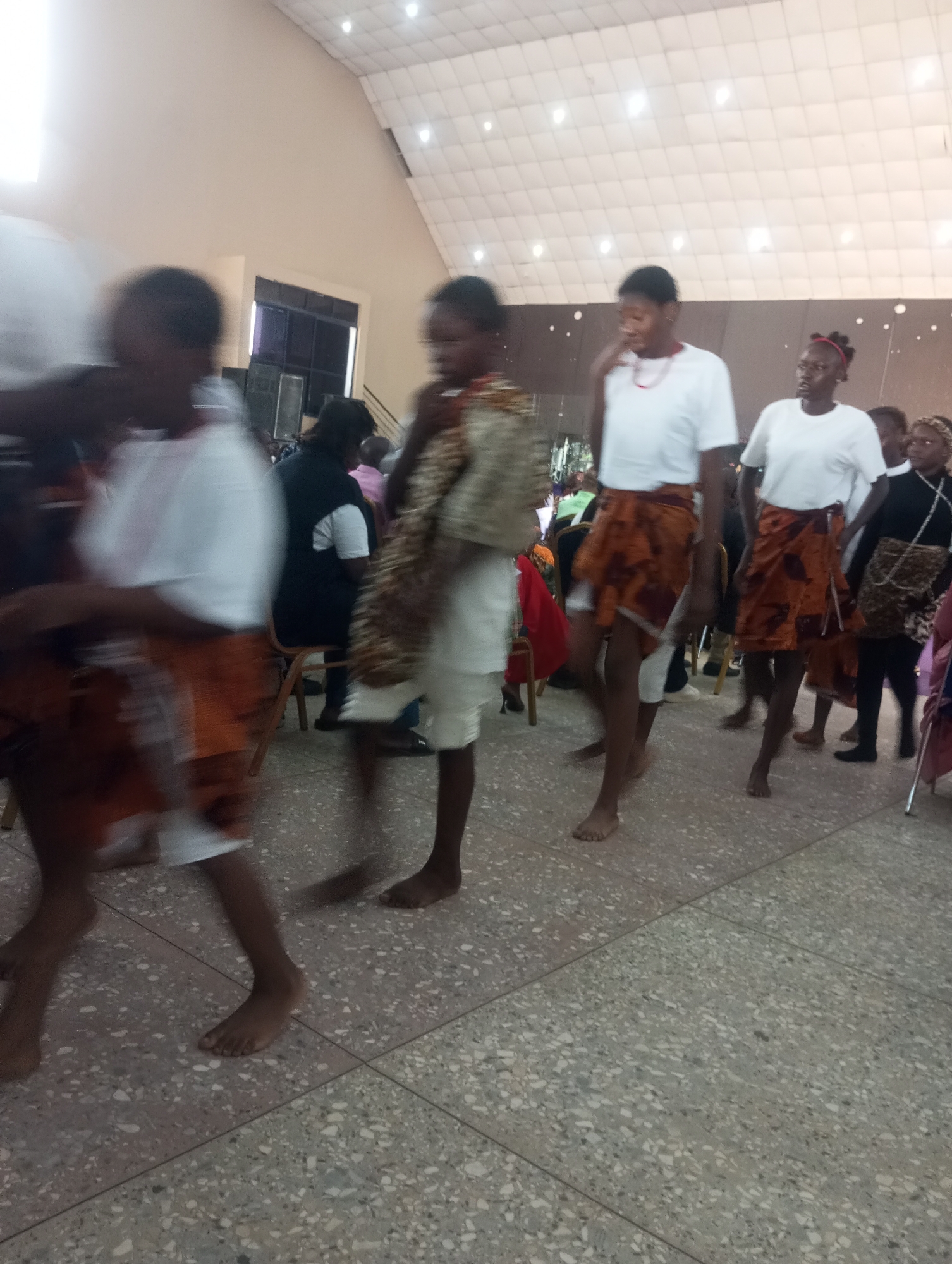In Gura Top, Nigeria, a group of individuals, predominantly women and girls, are captured in motion within an indoor venue, possibly a community hall or a church. The lighting suggests an artificial environment, with overhead lights illuminating a speckled floor and a patterned ceiling. The subjects are dressed in a combination of white t-shirts and patterned wrap-around skirts or cloths, with some wearing traditional attire. Their movements suggest they are dancing or participating in a procession. In the background, other individuals are visible, some seated and others standing, observing the activity. The overall scene depicts a cultural event or celebration taking place.