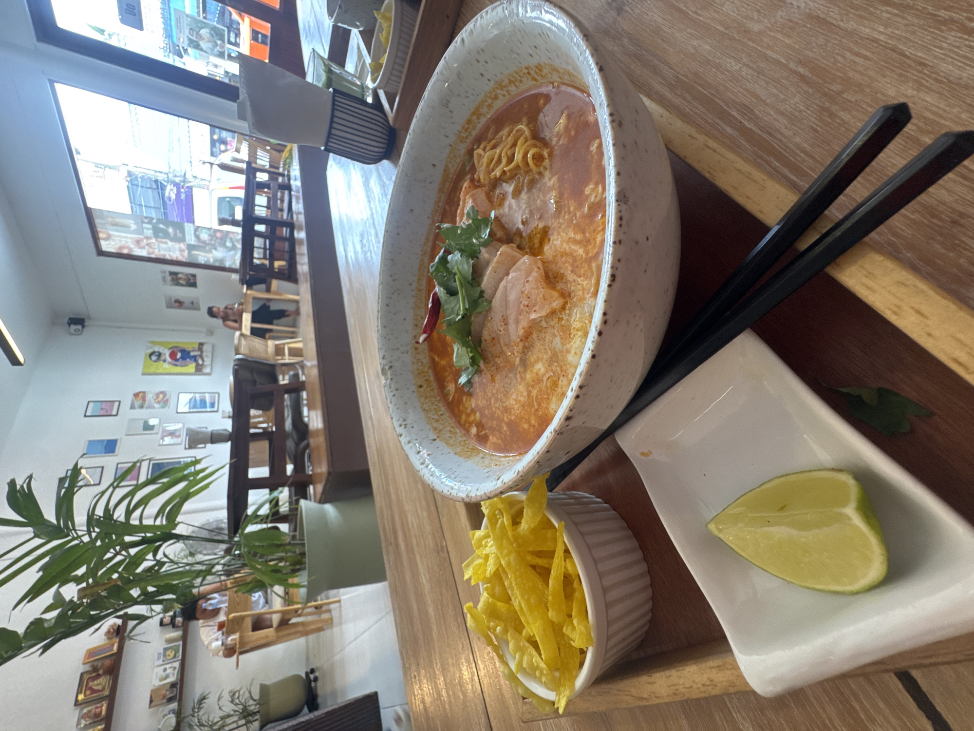 In a well-lit restaurant on Koh Phangan, Thailand, a bowl of Khao Soi sits prominently on a wooden table. The dish features noodles in a rich, creamy curry broth, topped with sliced meat, cilantro, and a dried chili. To the side, a small white ramekin holds crispy fried noodles, and a white rectangular dish contains a lime wedge. A pair of black chopsticks rests on the table beside the meal. In the background, a large potted plant with lush green leaves stands near a window. The restaurant's interior is visible, with other tables, chairs, and decorative artwork adorning the walls, suggesting a relaxed and inviting atmosphere.