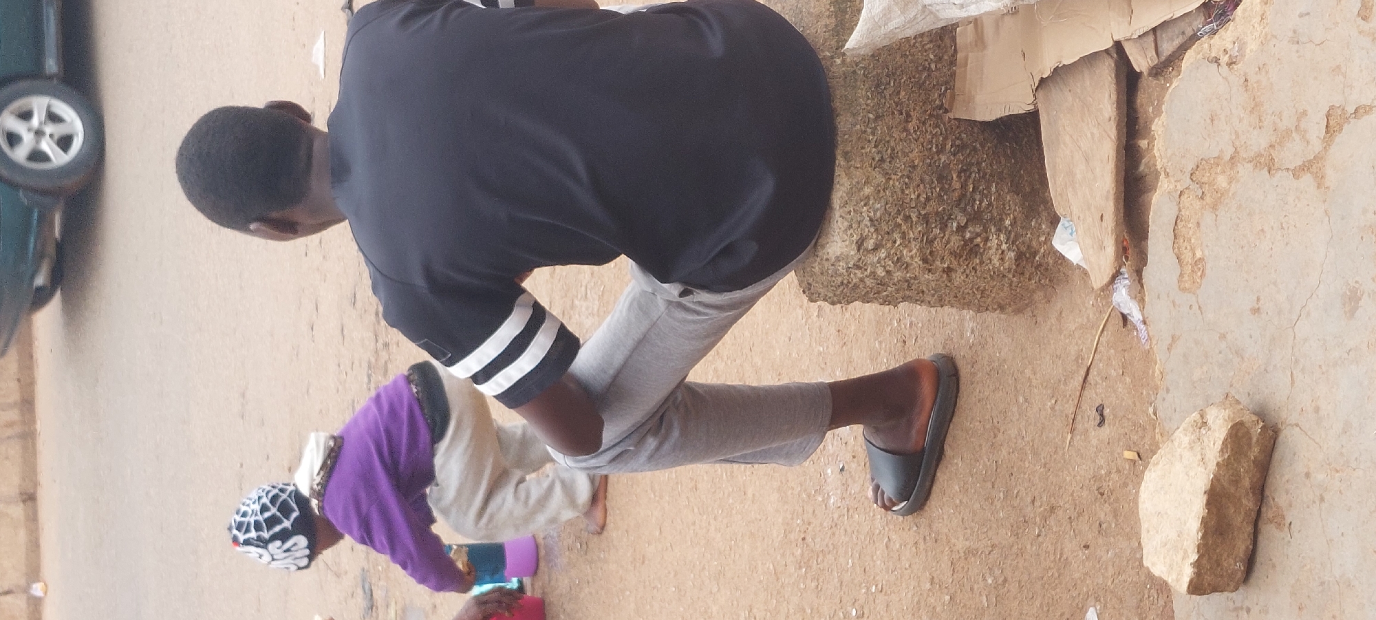 An outdoor scene depicts two individuals, an adult male and a child, on a dirt and partially paved surface.
In the foreground, an adult male, viewed from the back, is seated on a rough-hewn, light brown stone block. He wears a black short-sleeved shirt with three prominent white horizontal stripes on each sleeve, light grey sweatpants, and black slide sandals. His hair is dark and closely cropped.
To the left and slightly behind the adult, a child is observed from the back in a squatting position. The child wears a purple long-sleeved top or hoodie, light grey or tan pants, and a black and white beanie with a spiderweb pattern and partially visible white text that appears to read "ONS" or "SN." The child is barefoot and positioned near two small plastic containers, one blue and one purple, on the ground. A hand, likely belonging to another person, is visible on the far left edge of the frame, near the child.
The ground consists primarily of reddish-brown compacted dirt, interspersed with patches of cracked, lighter grey concrete or pavement in the immediate foreground. Scattered debris includes a small light brown rock, a dark twig, a small black object, white plastic fragments, and several pieces of cardboard, one large and folded.
In the background, a light-colored road is visible. Part of a dark green or blue vehicle with a silver five-spoke wheel is parked or moving on the road. A small yellow object is faintly discernible on the far side of the road. The lighting indicates daylight conditions.