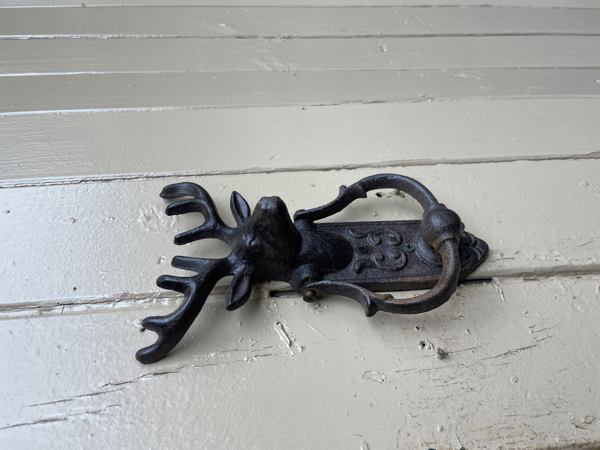 A black, cast-iron door knocker shaped like a deer's head is attached to a cream-colored wooden door in Da Lat, Vietnam. The deer's head is detailed with antlers and eyes, and a circular ring hangs from its mouth, designed to be used as the actual knocker. The door is composed of vertical planks, and the paint appears weathered, showing signs of age and wear. The background is slightly blurred, indicating a shallow depth of field, and the lighting suggests it is daytime.