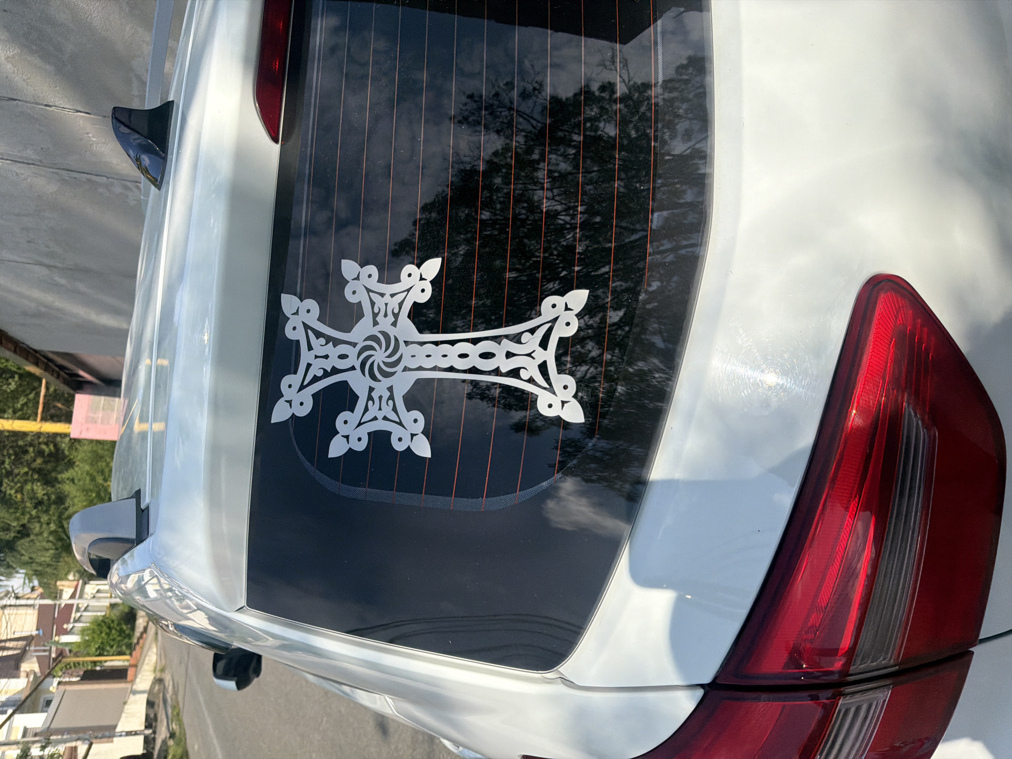 The image shows the rear of a white car parked on a street in Sochi, Russia. The car has a large, white sticker of an ornate cross on its rear window. The cross is decorative, with intricate swirling patterns in the center and petal-like extensions at the ends of its arms. The rear window of the car is tinted dark, and the vertical lines of the defroster are visible. A reflection of trees and the sky can be seen in the window. The car's red taillight is visible on the right side of the image. The street is paved, and a sidewalk with some buildings and greenery is visible in the background. The lighting suggests it is daytime, likely sunny, given the strong reflections on the car's surface. The scene appears to be a typical urban or suburban setting. There is no visible text in the image.