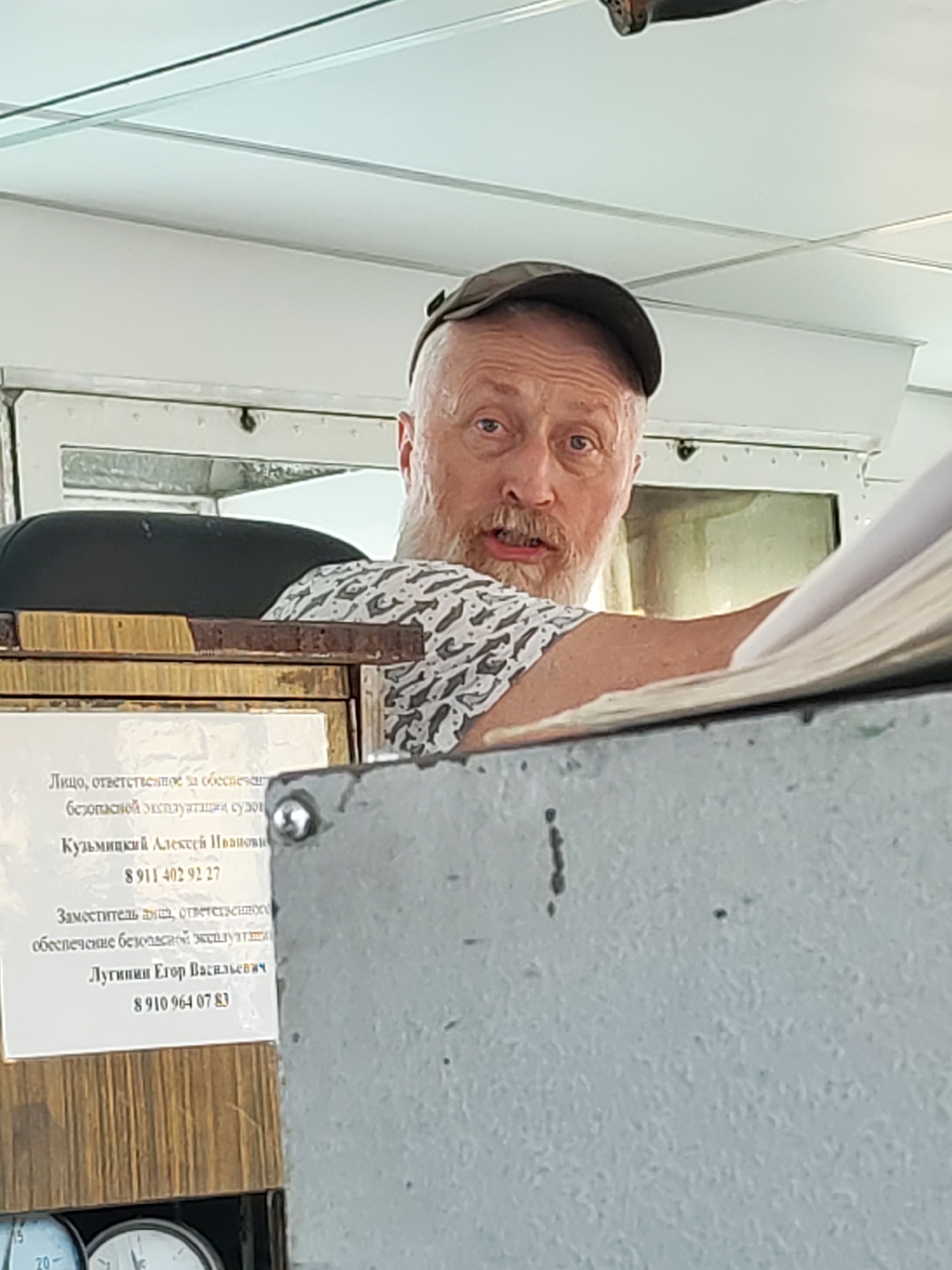 A medium shot captures a Caucasian man with a grey beard and a cap, partially obscured behind a grey metallic surface. The man is looking directly at the camera with a slight frown. Behind him, a white piece of paper is affixed to a wooden surface, with text in Russian. The Russian text translates to: "Person responsible for ensuring the safe operation of ships: Kuzmitsky Alexey Ivanovich 8911 402 92 27 Deputy commander responsible for ensuring safe operation: Luginin Egor Vasilyevich 8910 964 07 83". Based on the Russian text, the scene may be located in Russia, possibly the city of Yakhroma, Russia. The wooden surface has two gauges below the paper. The environment seems to be indoors, potentially within a control room or navigational area, possibly on a ship, in Yakhroma, Russia.
