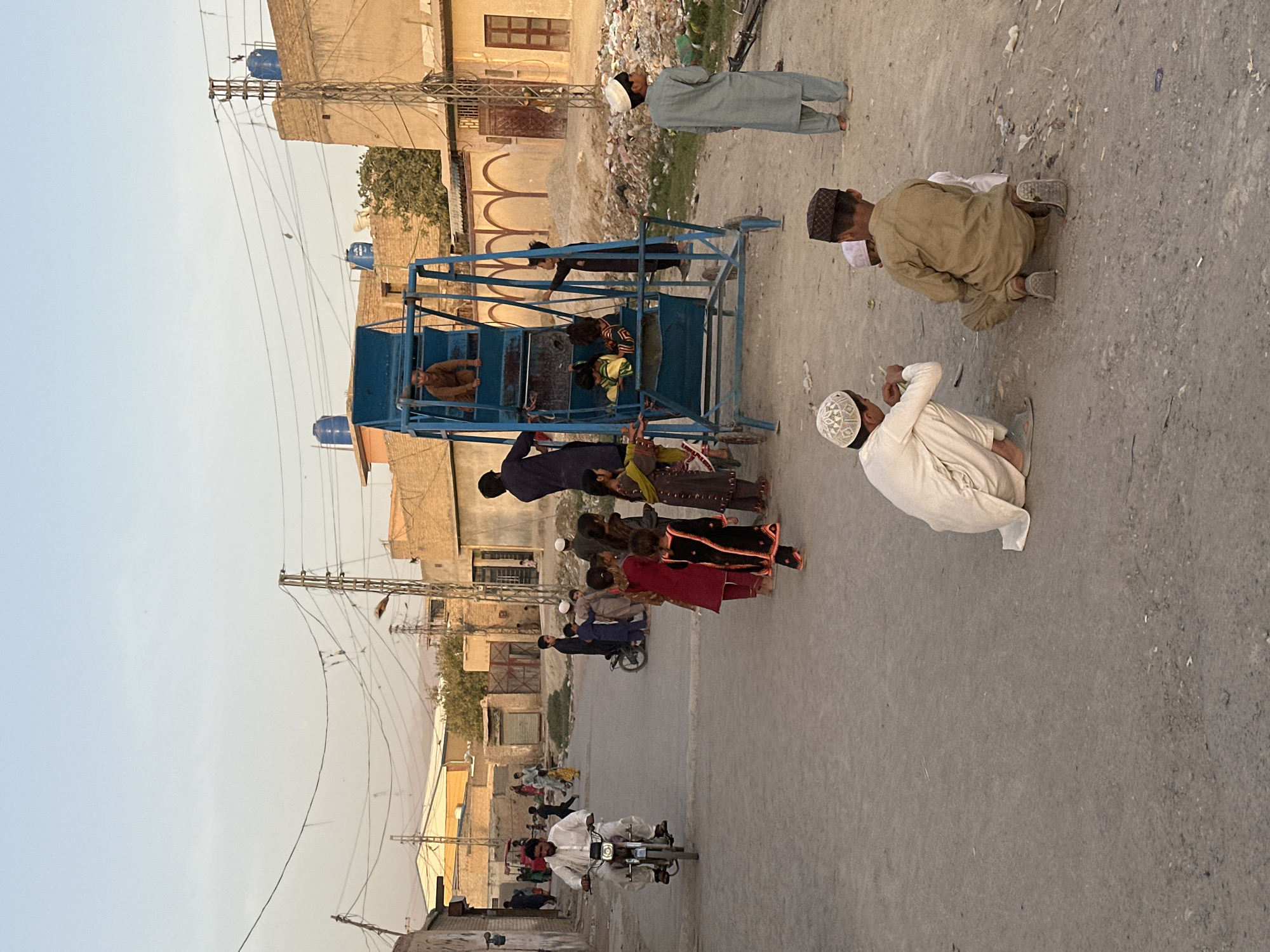 The image depicts a dusty street scene in Quetta City Tehsil, Pakistan, during daylight hours. In the foreground, two young boys are crouching on the ground. One boy, wearing a white cap and traditional clothing, is looking to the left. The other boy, in a brown outfit, is facing away from the camera. Behind them, a group of children, mostly girls dressed in colorful traditional attire, are gathered near a blue metal swing set. Some children are on the swing set, while others stand nearby. A young boy in a grey outfit stands further to the right, looking towards the left. In the mid-ground, a man is riding a white motorcycle towards the left of the frame. Several other figures, including children and adults, are scattered throughout the street, some walking, others standing. The background shows low-rise buildings constructed from mud brick and cement, with some evidence of disrepair. Power lines are visible crisscrossing overhead. The ground is unpaved and dusty, with some scattered debris. The overall impression is of a community gathering on a street with limited infrastructure.
