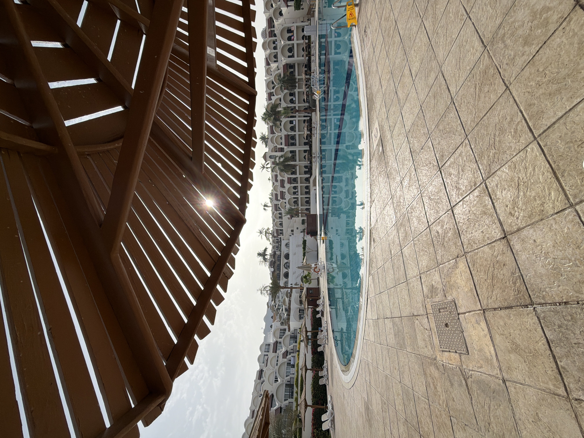 The image shows a view of a swimming pool area at a resort in Sharm El Sheikh, Egypt. The photo is taken from under a wooden pergola or shade structure. The pool is edged with light brown patterned tiles. White multi-story hotel buildings are visible in the background, reflecting in the pool's water. Palm trees dot the scene between the buildings. In the foreground, a manhole cover is visible on the tiled area. The sky is overcast. The sun is positioned behind the slats of the pergola, creating a starburst effect.