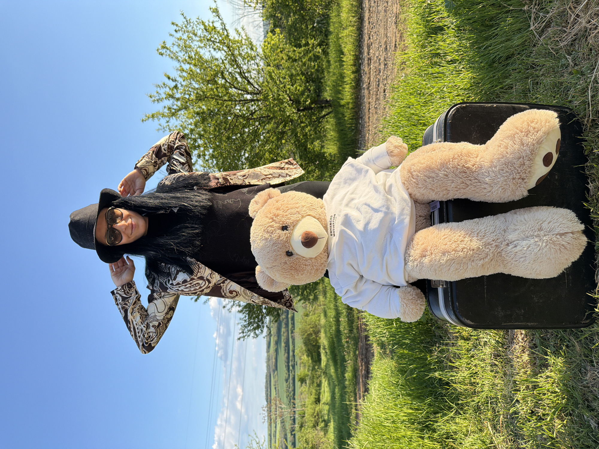 A woman wearing a black hat, sunglasses, a black shirt, and a patterned jacket is posing next to a large teddy bear wearing a white shirt. The teddy bear is lying on a black suitcase. They are in a grassy field with a tree in the background and a view of the countryside. The location is near Pleven, Bulgaria.