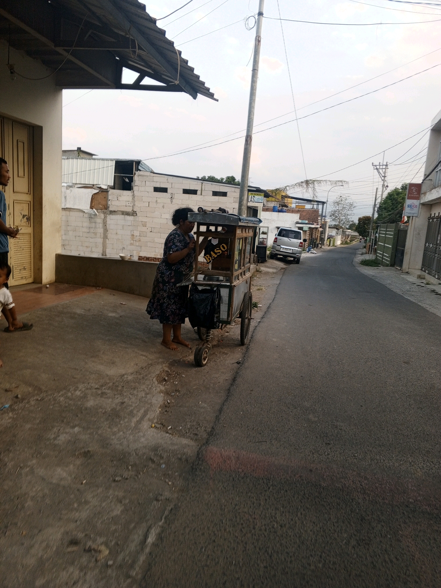 A medium shot captures an outdoor street scene with a woman standing by a food cart. The woman, wearing a dark floral dress, is barefoot and appears to be tending to the cart. The cart is wooden with glass panels and has "BASO" written on it. A child in a white shirt and dark shorts is visible on the left, and a man in a blue shirt is partially visible behind the child. The environment is a street with a sidewalk, lined with buildings and a road that curves into the distance. Electrical wires crisscross the sky, and a silver van is parked on the side of the road. The time of day appears to be late afternoon or early evening, indicated by the lighting and the soft sky.