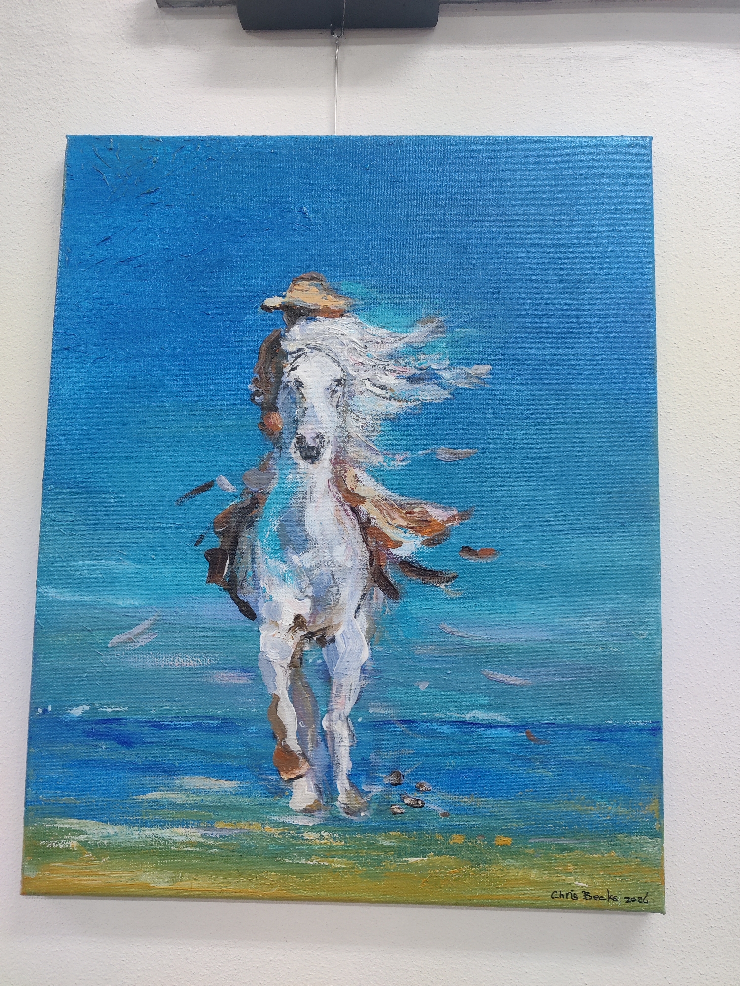 A painting of a white horse with a rider on its back, galloping through shallow water. The horse's mane is flowing in the wind. The rider is wearing a hat and a light-colored shirt. The background is a bright blue sky and water. The painting is signed "Chris Beeks 2026" in the lower right corner. The painting is hanging on a white wall, possibly in a gallery or home.