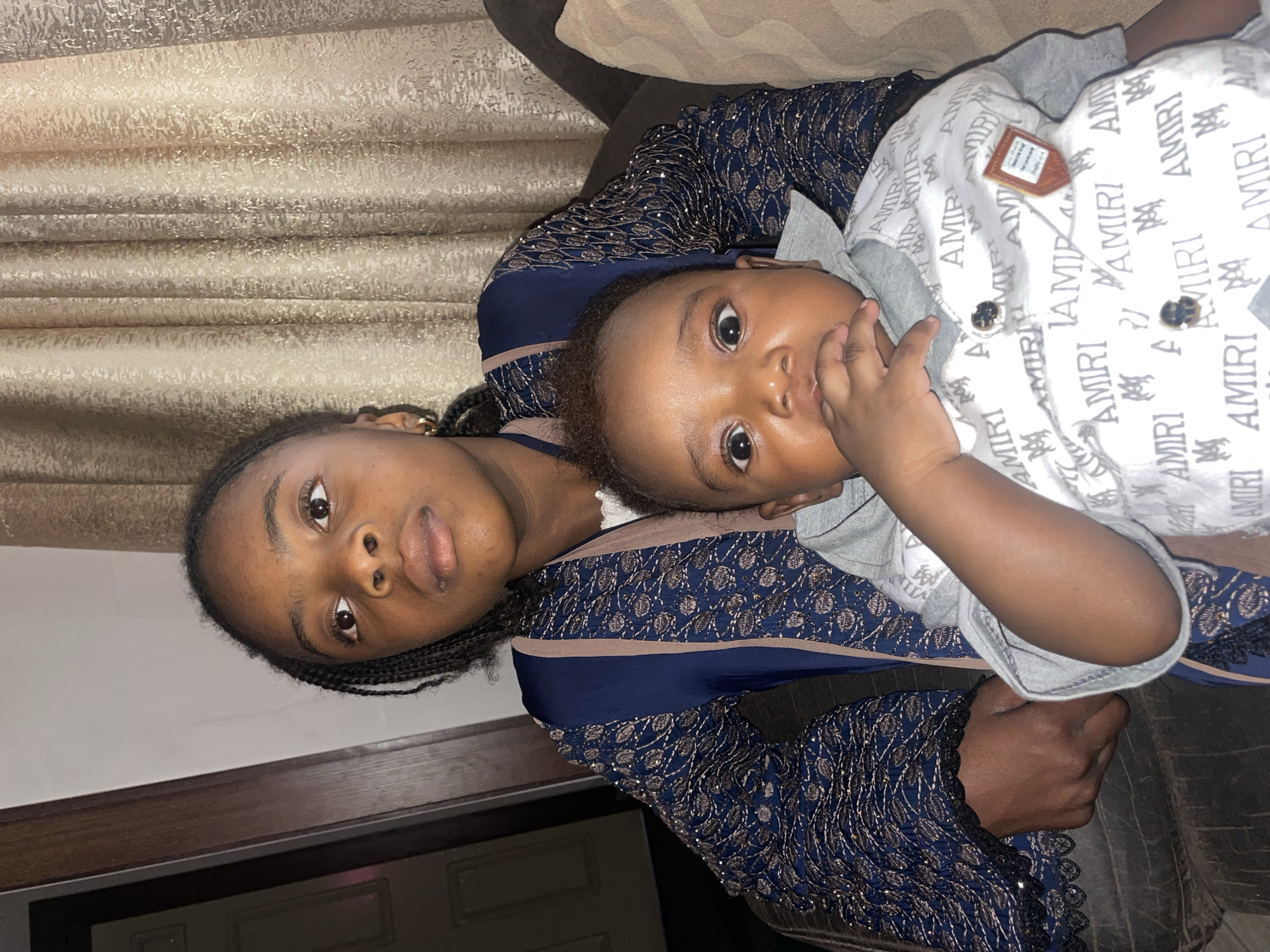 A medium shot captures a young woman and an infant lying down. The woman is positioned on the left, and the infant is resting on her chest and arm. The woman has dark skin, full lips, and dark eyes that are directly gazing at the camera. Her hair is braided. She is wearing a navy blue patterned dress. The infant also has dark skin and dark eyes that are looking at the camera. The infant's right hand is near its mouth. The infant is wearing a light gray shirt under a white button-down shirt that has "AMIRI" printed all over it. The background includes a wall, a curtain, and a sofa. The location is likely indoors, but not identifiable as Jos, Nigeria based on the visual information.