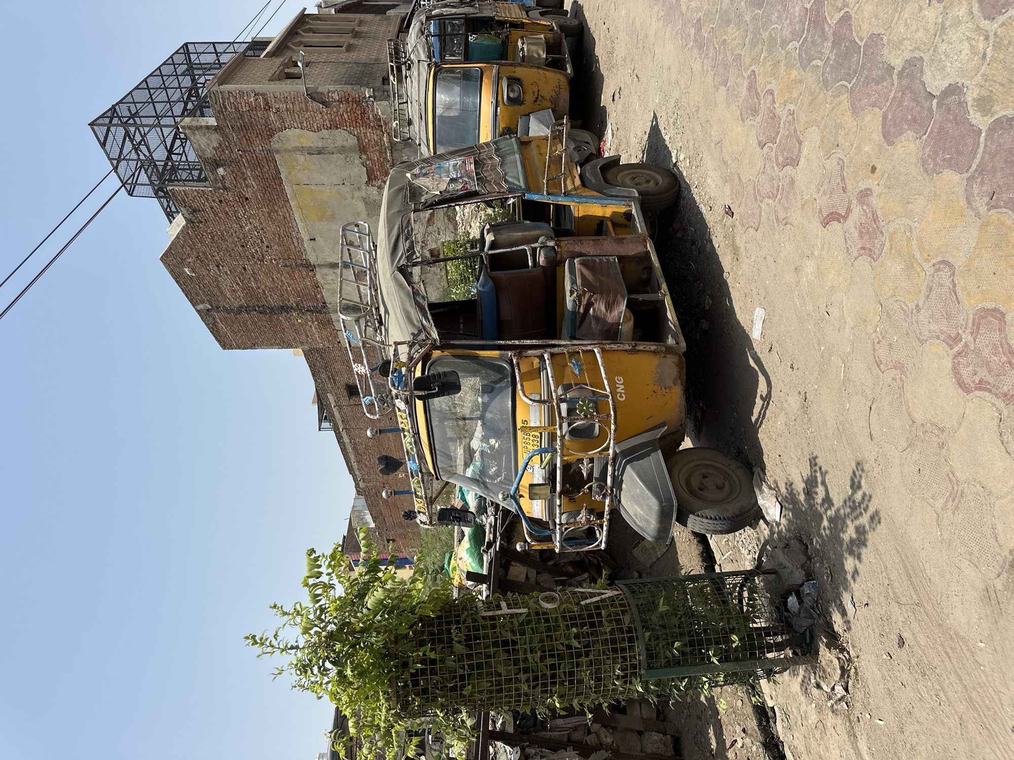 In Vrindavan, India, a yellow auto-rickshaw sits idle on a paved street. The vehicle, weathered and showing signs of age, is partially obscured by a large, round, wire-mesh trash bin containing refuse and some sparse greenery. Behind the auto-rickshaw stands an aged brick building, with a metal cage-like structure attached to its upper reaches. The sky is a clear blue. The street paving features an interlocking pattern of red and tan blocks. Another auto-rickshaw is partially visible behind the first.