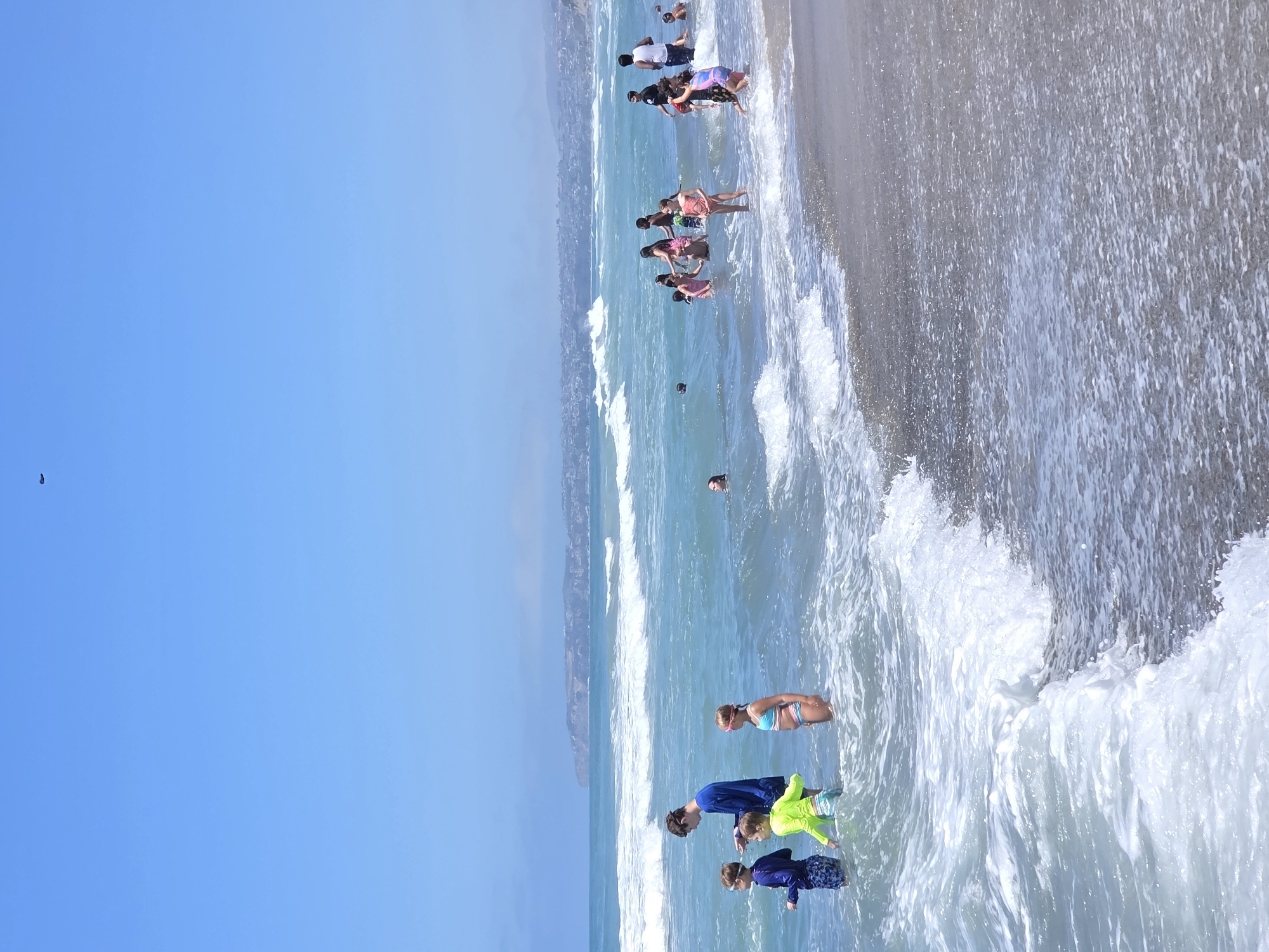The image depicts a sunny day at a beach in San Clemente, California. Several people, predominantly children, are in the ocean wading and playing in the waves. In the foreground, a group of children are standing in the shallow water. To the left, a young boy in a blue rash guard and patterned swim trunks stands with a person in a blue shirt. Next to him is another boy in a bright yellow rash guard and green swim trunks. A girl in a blue bikini top and patterned bottoms, wearing pink goggles, stands to the right of the boys. In the mid-ground, more people are scattered in the water, some swimming and others standing. Further in the background, the coastline of San Clemente is visible with houses and hills. The ocean is a clear blue-green with white-capped waves rolling towards the shore. The sky is a bright, cloudless blue. A small dark object is visible in the upper center of the sky.