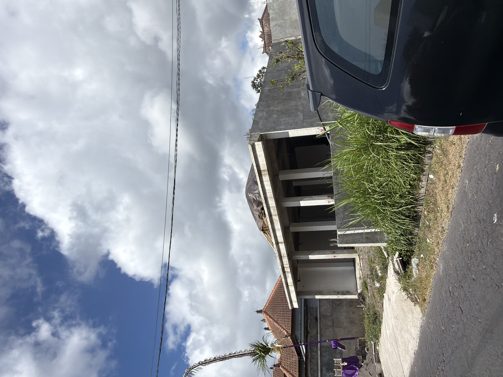 A black minivan is parked on the right side of a paved road. In front of the minivan and to the left, a single-story house is under construction. The house is made of concrete and has an open roof covered with a tarp. Tall green grass is growing in front of the house. To the left of the house, there is another house with a red tile roof. A tall, decorative Balinese banner made of palm fronds and purple fabric is planted in the ground next to the house with the red roof. The sky is cloudy with patches of blue. The scene appears to be in Kintamani, Indonesia.