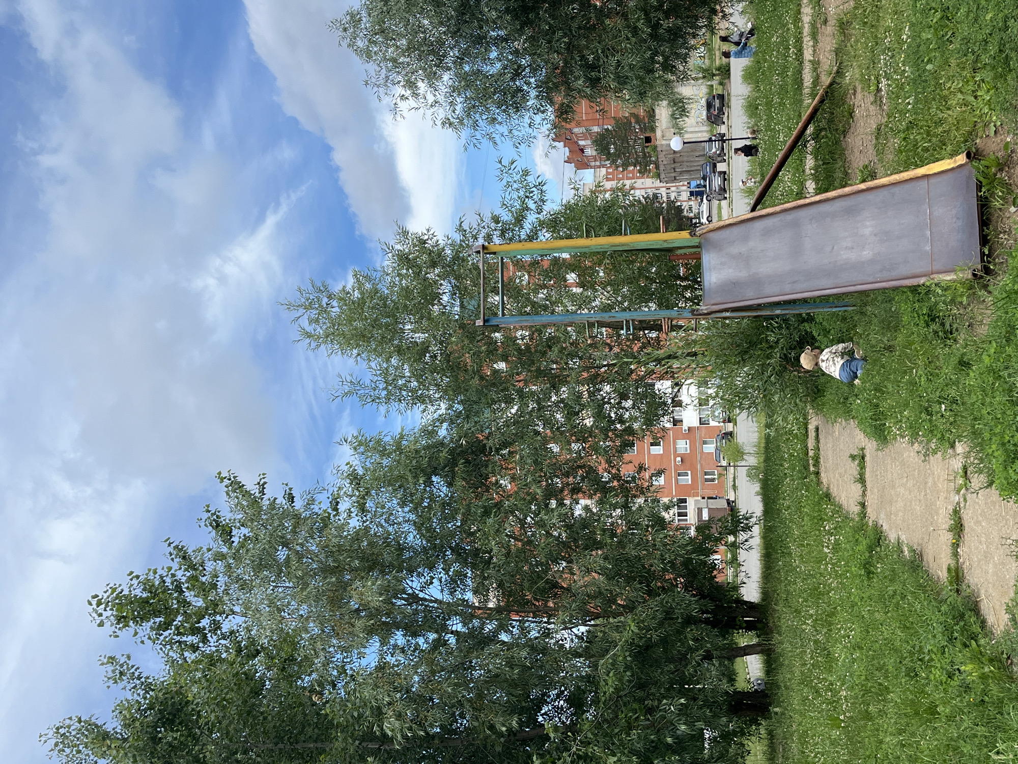 The image depicts a playground scene in the city of Chaikovsky, Russia, on a cloudy day. A metal slide with yellow and blue painted supports is prominent in the foreground, with a rusty brown support pole extending from its side. A young child, wearing a beige hat and patterned shirt, is crawling on the grass near the bottom of the slide. A concrete path meanders through the grassy area, dotted with small white flowers. Several trees with lush green foliage are scattered throughout the scene, partially obscuring apartment buildings in the background. The buildings are constructed of brick and concrete, with numerous windows visible. In the distance, cars are parked along a road, and a few individuals are seated on a bench. The sky is a mix of blue and white clouds.