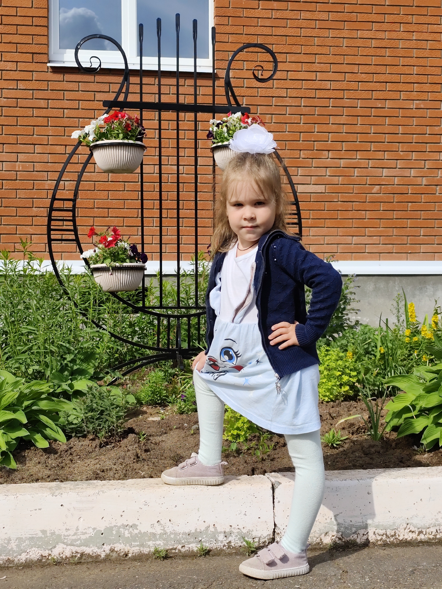A young girl, likely a toddler or preschooler, confidently poses outdoors in what appears to be a garden or yard in Фоки, Russia. She stands with her left foot elevated onto a low, light-colored concrete curb, her knee bent, and her right hand resting on her hip, looking directly at the camera with a serious expression.
The girl has light-colored hair, styled with a prominent white flower or bow on the top of her head. She is dressed in a light blue tunic or dress featuring a large cartoon character's face with big blue eyes on the front, worn over pale green or light blue leggings. She completes her outfit with a dark blue, textured, zippered cardigan and light pink or beige sneakers with velcro straps.
Behind the girl, a decorative black metal trellis or archway with ornate scrollwork at its peak features three white potted plants, each overflowing with a mix of red, white, and purple flowers. Further in the background, a reddish-brown brick wall, part of a building, is visible, along with a white-framed window reflecting a partly cloudy sky. A garden bed filled with various green plants and some scattered yellow flowers lines the area behind the concrete curb. The foreground features a dark, paved surface. The bright, clear lighting indicates a sunny daytime setting, likely mid-morning or afternoon. No text is visible in the image.
