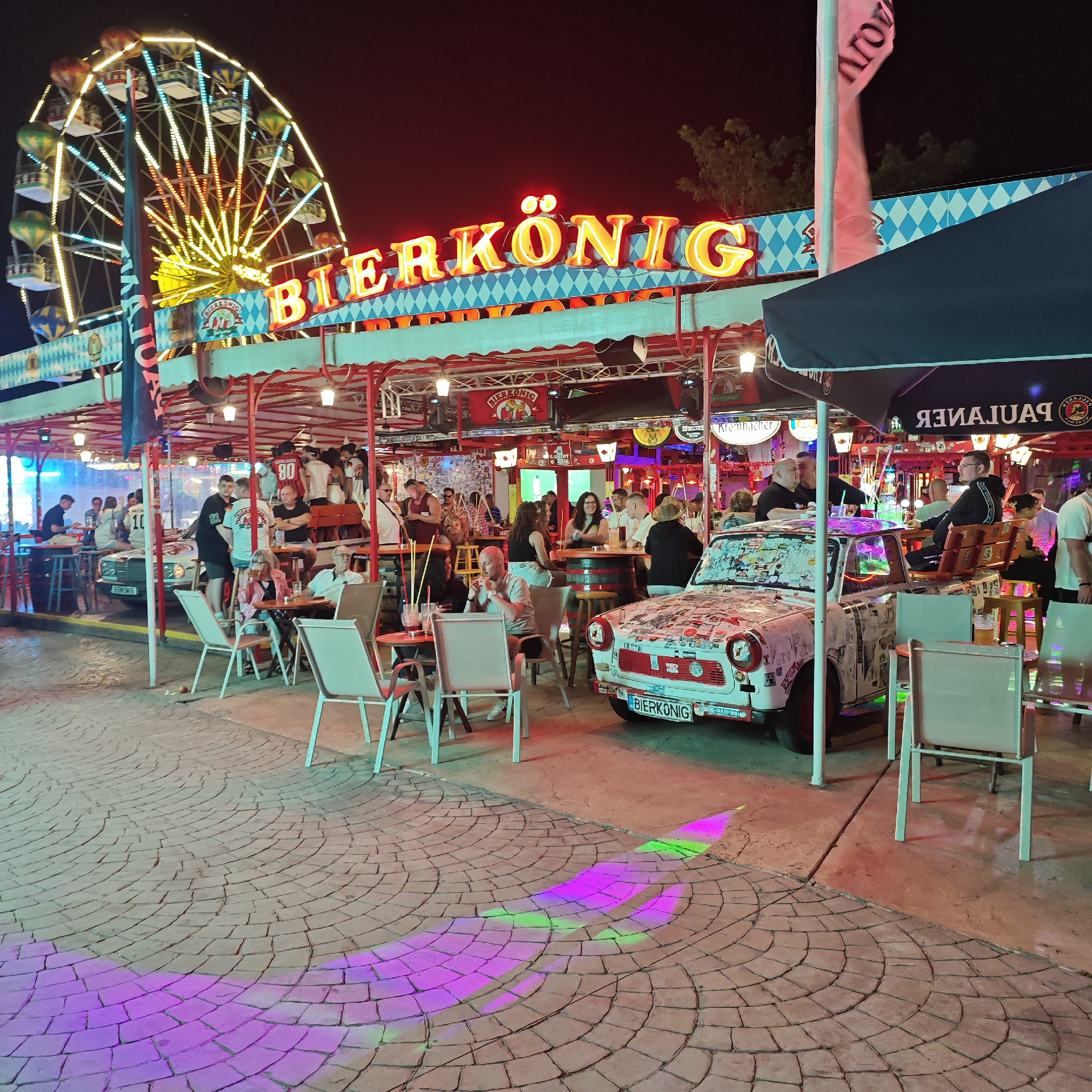 This image depicts a lively outdoor bar area at night, likely located in Varna, Bulgaria, as suggested by external context. The scene is illuminated by neon signs and string lights, with a Ferris wheel visible in the background. A prominent sign with "BIERKONIG" in glowing orange letters adorns the establishment. Patrons are seated at tables, some engaged in conversation while others are being served drinks. A vintage car, covered in stickers, is prominently displayed as a decorative element, with a bench fitted to its roof. The ground is paved with interlocking stones, and colorful lights are projected onto the ground, creating a festive atmosphere. The overall impression is one of a bustling entertainment venue.