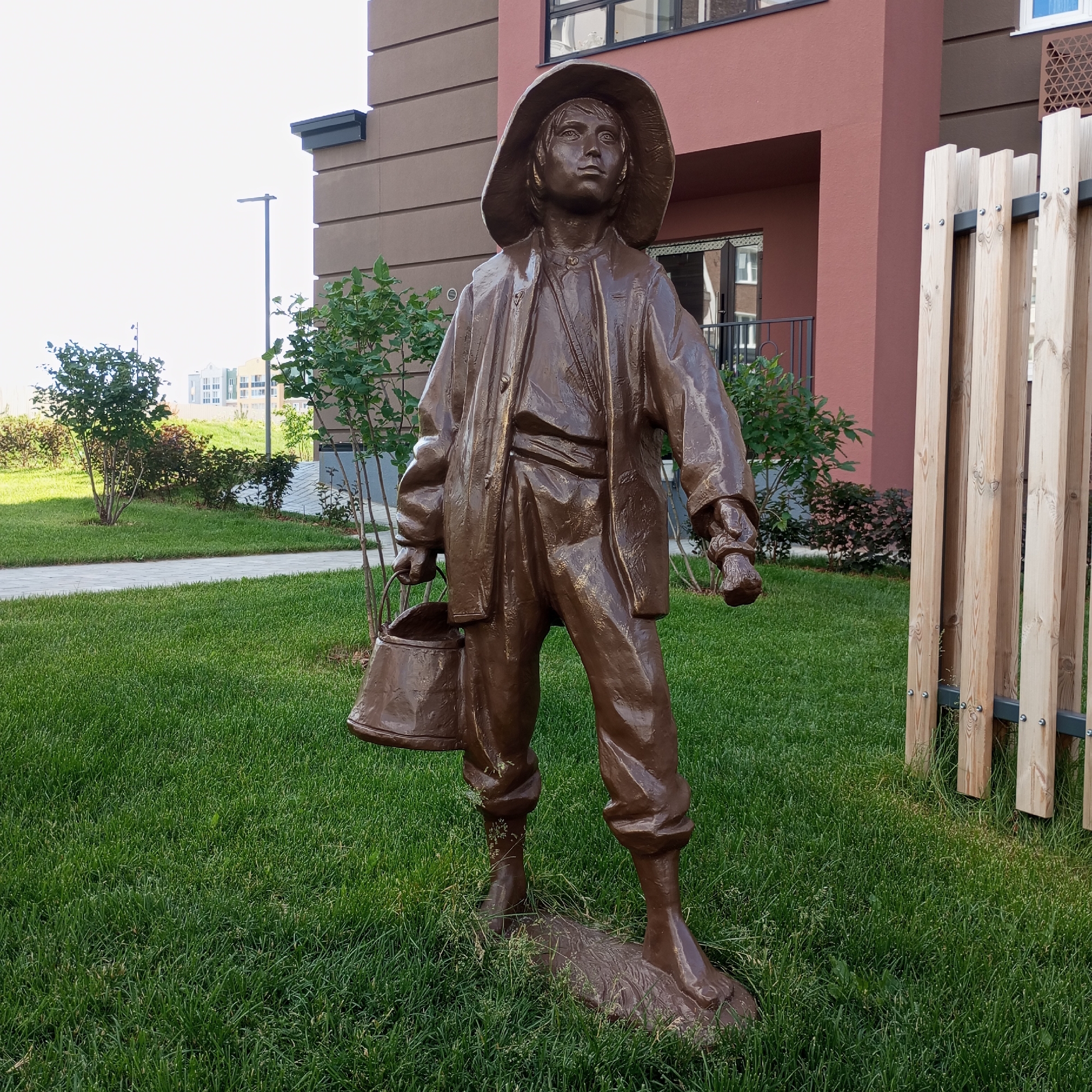 A bronze statue of a young person in a wide-brimmed hat and loose-fitting clothing stands on a patch of green grass. The figure is holding a bucket in their left hand and their right hand is clenched. The statue is positioned in front of a modern building with a red and brown facade. To the right of the statue is a wooden fence made of vertical planks. In the background, there are more buildings and lush green trees, suggesting an outdoor setting in a well-maintained area. The lighting indicates it is daytime, likely with clear or partly cloudy weather, casting a soft shadow from the statue onto the grass. The overall scene appears to be in a residential or public space, given the presence of the building, fence, and manicured lawn. No visible text is present in the image. The statue's expression is contemplative as they look upwards.