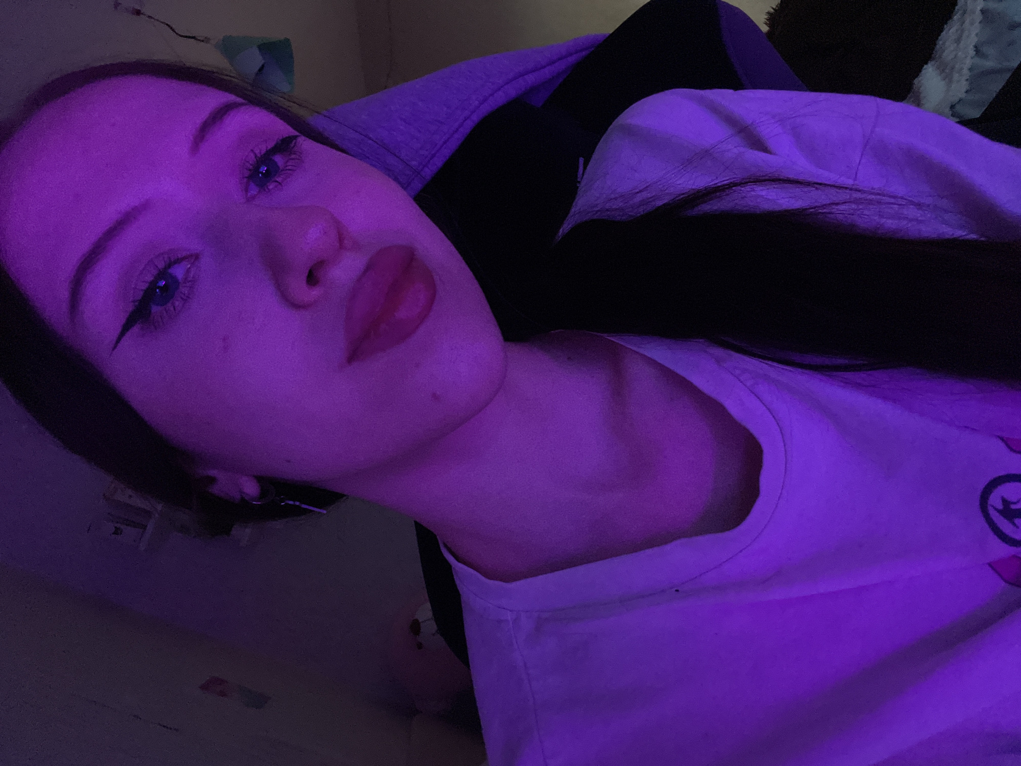 The image is a close-up, selfie-style shot of a young woman illuminated by purple lighting. She is wearing a white t-shirt and has dark hair. Her makeup includes winged eyeliner and lipstick. The background is out of focus but appears to be an indoor setting with some shelving and objects visible. There are no discernible elements in the image that would suggest a specific location within Votkinsk, Russia, or any other city.