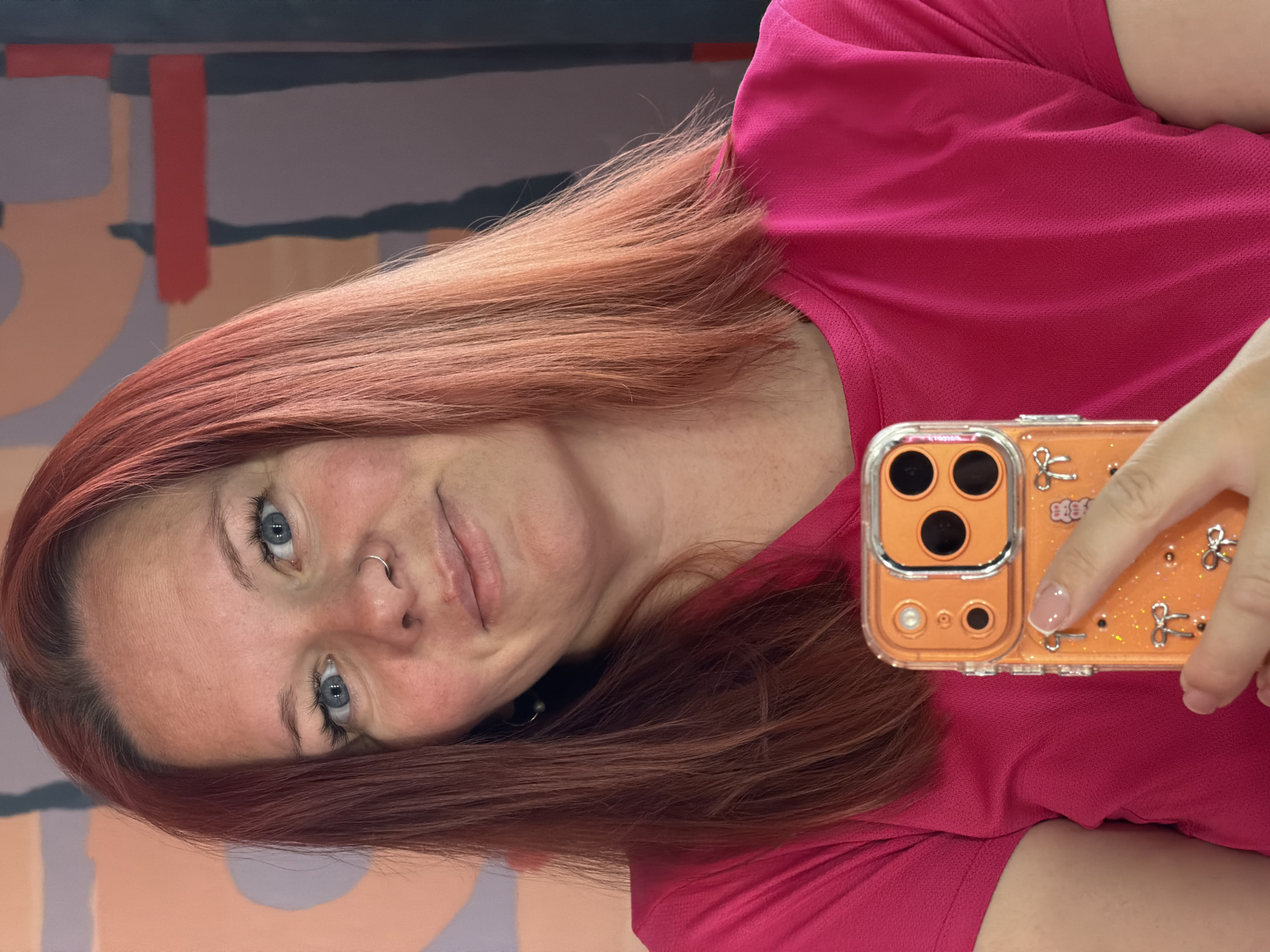A woman is taking a selfie with her orange phone. She is wearing a pink shirt. She has red hair and blue eyes. She has a nose ring. The background is a wall with abstract art. I cannot determine the location in the city of Chaikovsky, Russia.