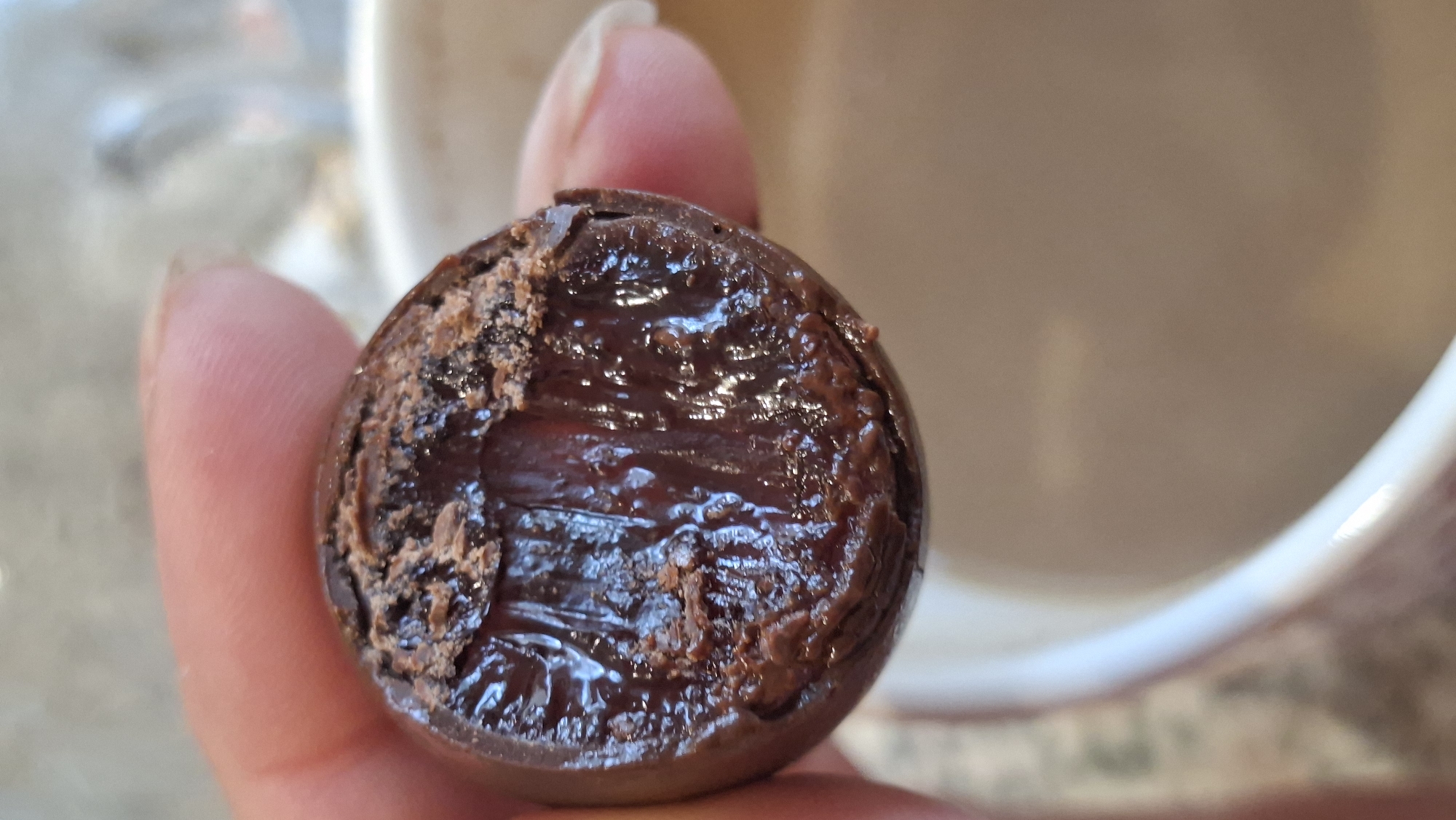 A close-up, top-down view shows a person's fingers holding a dark brown, round chocolate confection that has been bitten into, revealing a glistening, gooey, dark red filling. The chocolate shell is partially crumbled around the bitten edge, with fine brown specks visible. The background is blurred, showing the rim of a white mug containing a light brown beverage, likely coffee or hot chocolate, and a mottled surface, possibly a table. The image's setting is indeterminate and cannot be linked to Myadzyel, Belarus.