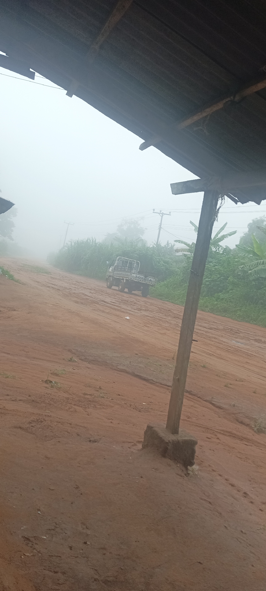 The image shows a dirt road in Amauzu, Nigeria, shrouded in heavy fog. A Toyota pickup truck is driving up the road. Lush, green vegetation lines the roadside. Utility poles and power lines are visible in the background. In the foreground, a wooden post with a concrete base stands under the edge of a corrugated metal roof. The ground is red dirt.