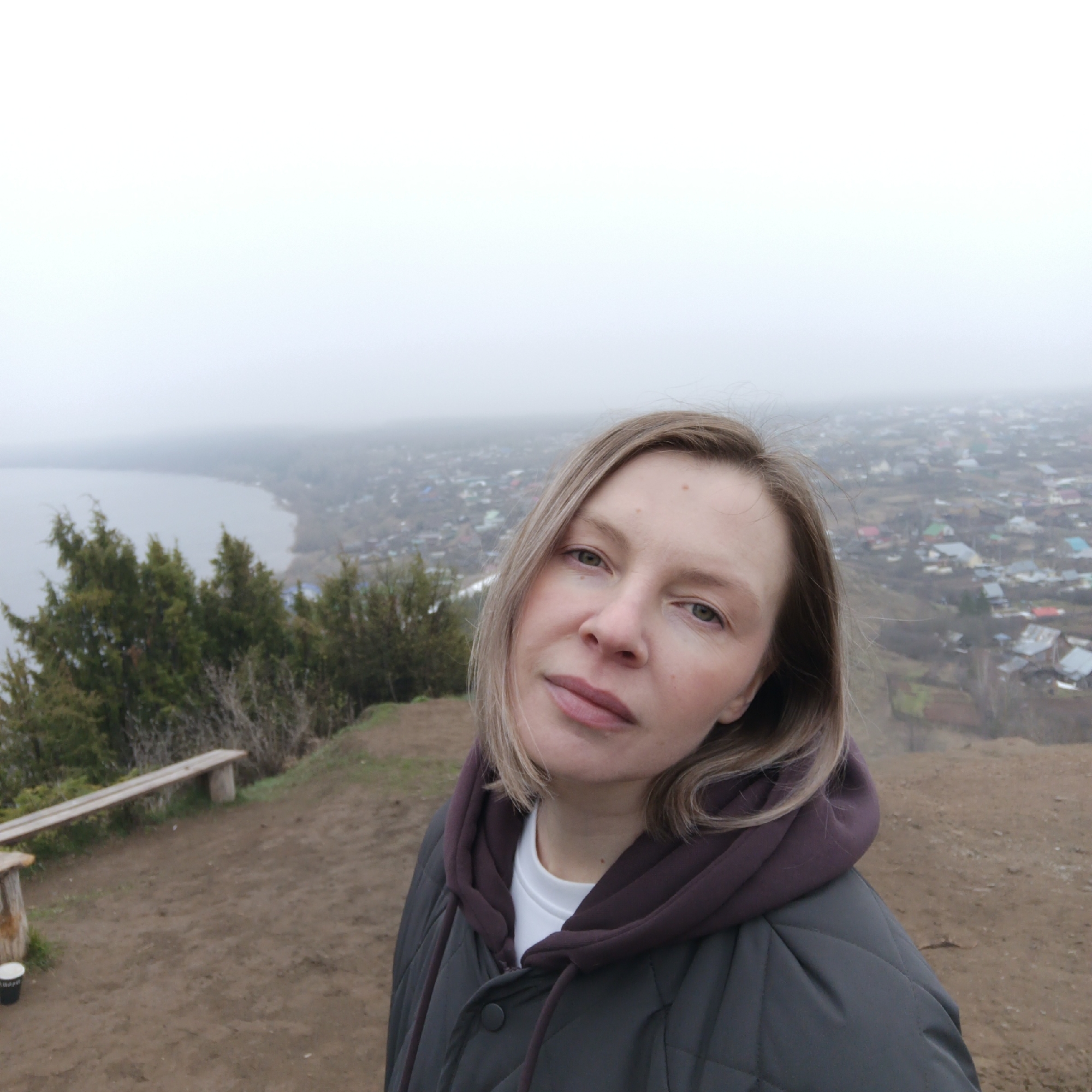 The image is a selfie of a fair-skinned woman with short blonde hair and green eyes. She is wearing a white t-shirt and a grey quilted jacket with a purple hoodie underneath.
The background reveals a foggy cityscape, suggesting a high vantage point. A body of water borders the town. To the left of the woman are green trees and a simple wooden bench. The ground is dirt and foliage. There is a cup on the ground near the bench.
Based on the cityscape visible and the information provided, the location is Ольховка, Russia. The person is likely standing on a hill overlooking the town.