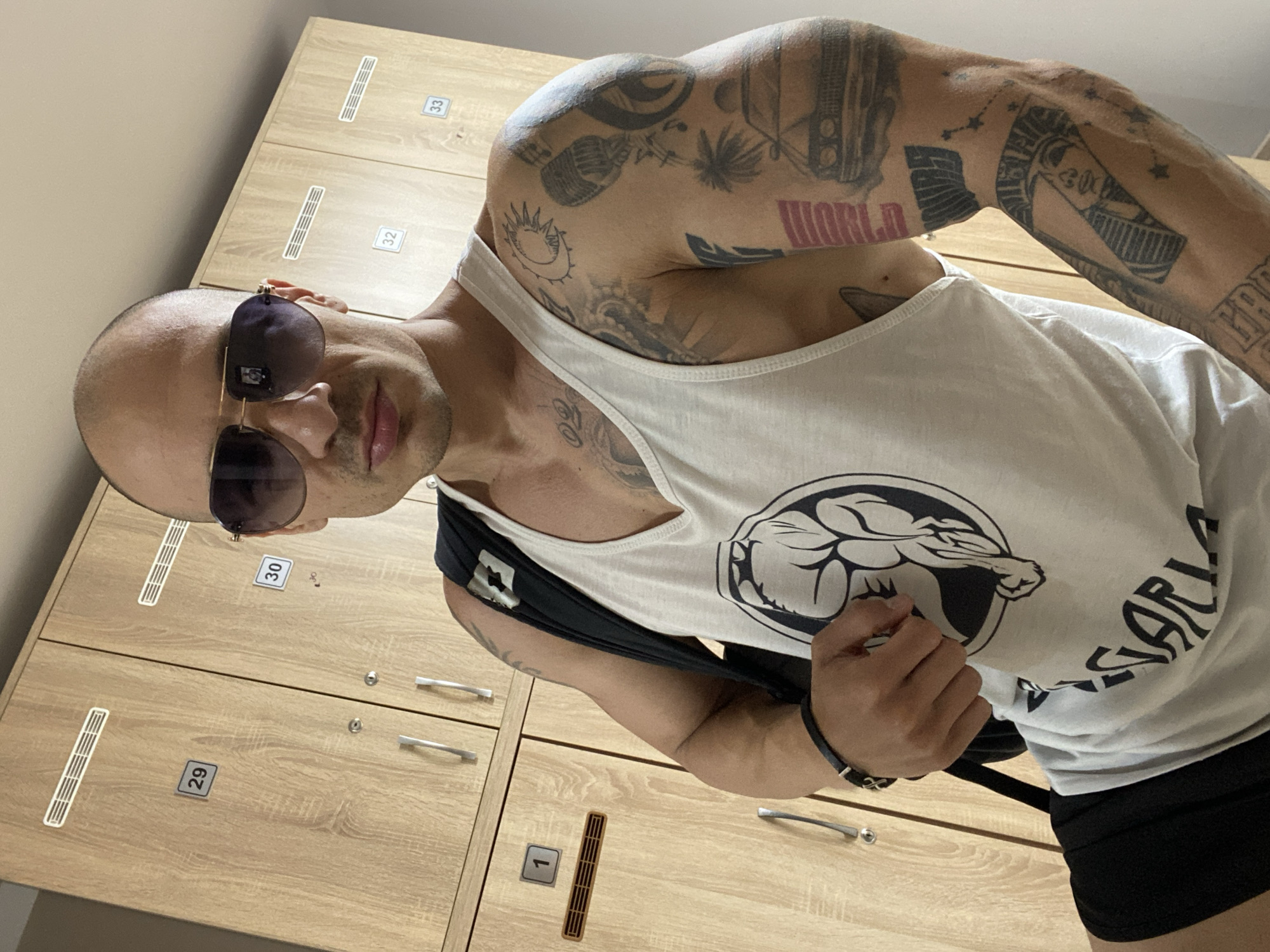 A bald, muscular man with tattoos on his arms poses indoors, wearing a white tank top, black shorts, and sunglasses. He is in what appears to be a locker room, with light wood-toned lockers visible behind him, numbered from 1 to 33. The exact location within the city of Pleven, Bulgaria is indeterminable from the image. The man appears to be holding a black bag strap.