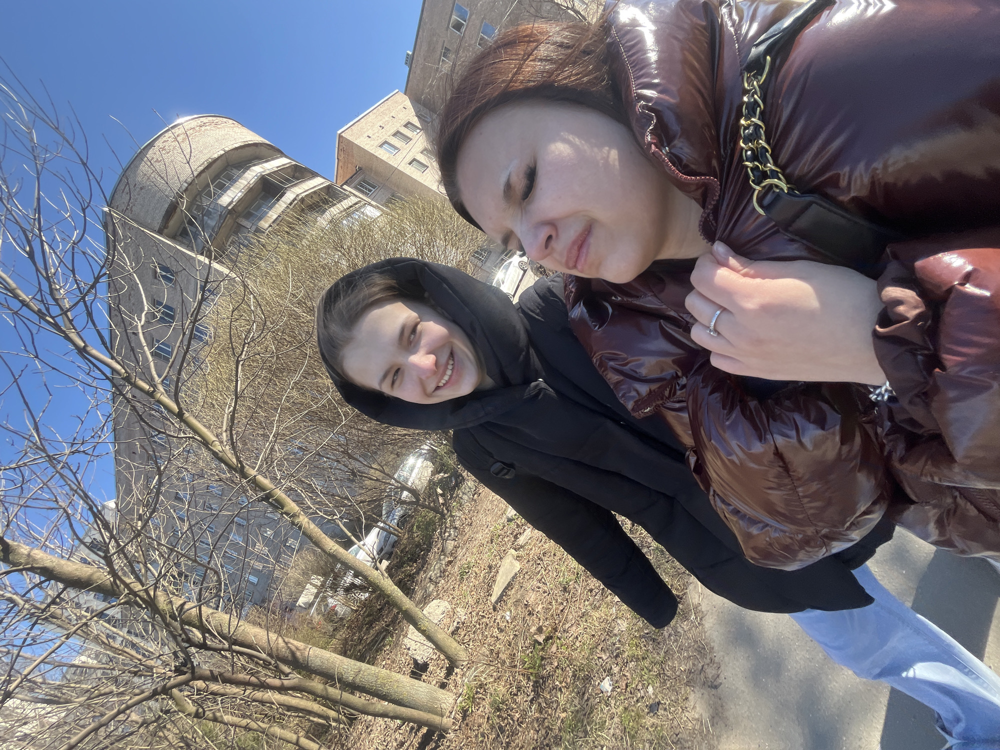 Two women pose for a selfie outdoors. One woman wears a black hooded jacket, and the other wears a shiny brown puffer jacket. They stand on a paved area next to bare trees and a grassy patch. Buildings are visible in the background. The city of Saint Petersburg, Russia, is identified as the location.