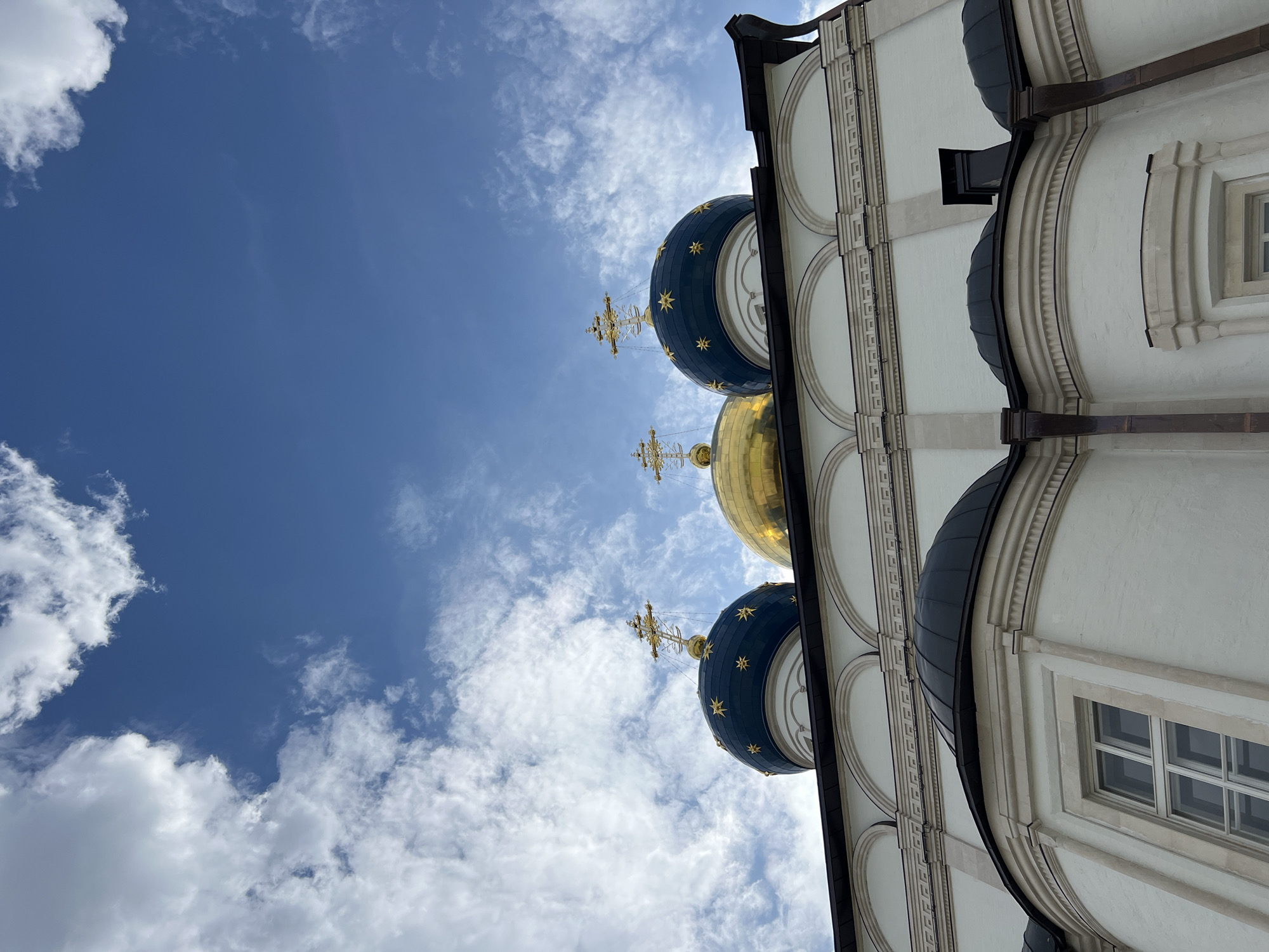 The image depicts the domes of the Resurrection Cathedral in Tver, Russia, against a blue sky with white clouds. Three prominent domes are visible: two dark blue domes adorned with gold stars and a central, golden dome. Each dome is topped with an ornate gold cross. The lower portion of the image shows the white facade of the cathedral, featuring architectural details like arched windows and decorative moldings. The style of the architecture suggests a Russian Orthodox church. The lighting indicates it is daytime, likely with sunlight illuminating the scene. No people are visible in the frame.