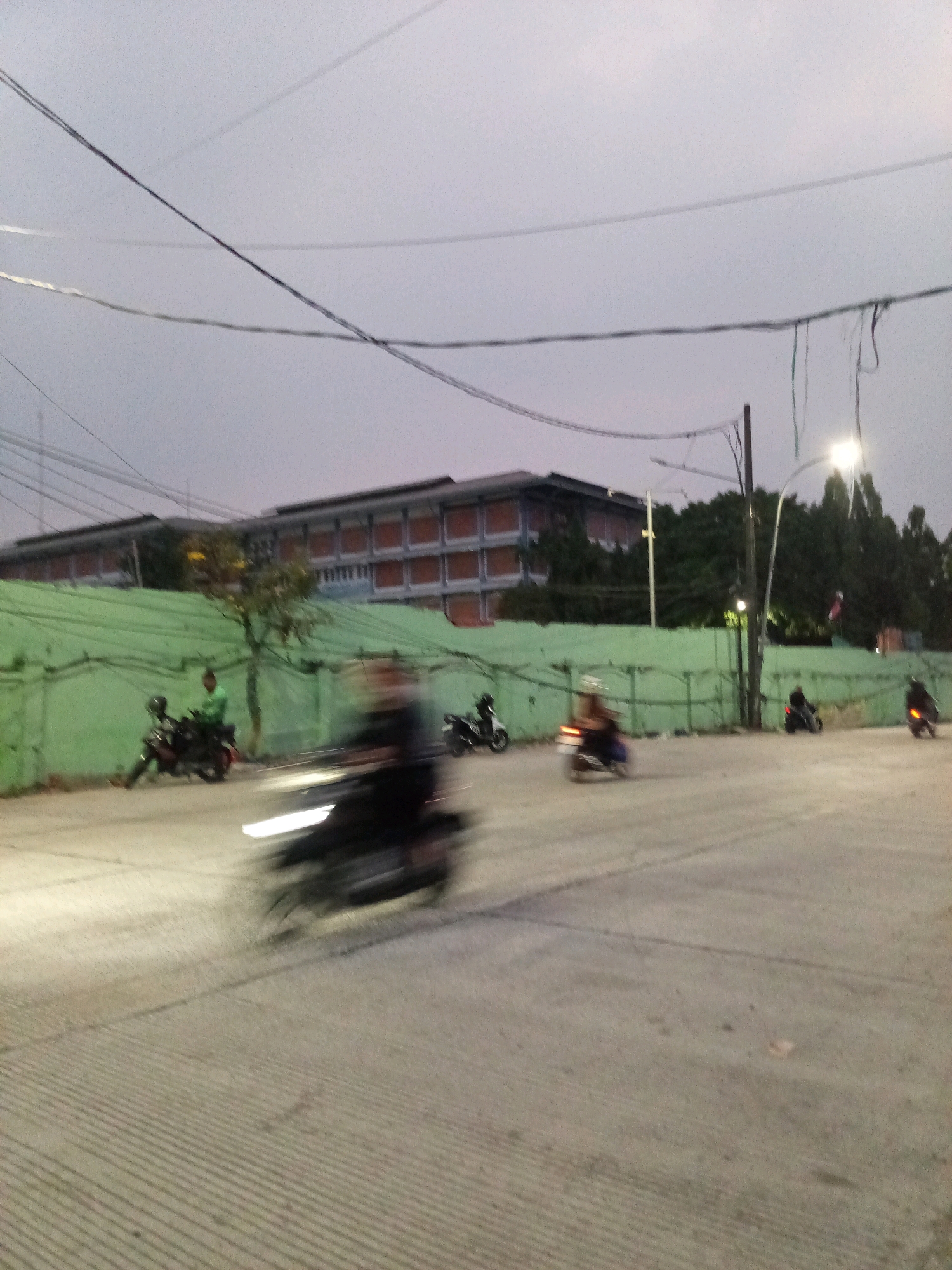 The image captures a street scene at dusk in Bekasi, Indonesia, likely near a school or institutional building. In the foreground, the pavement has a textured, ribbed pattern. Several motorcycles are in motion, their forms blurred by the low light and movement, suggesting a busy street. One motorcycle, closer to the viewer, is a dominant dark blur with bright white headlights.
In the middle ground, a person in a green shirt is standing next to a parked motorcycle. Behind them is a light green, textured wall or fence, which stretches across the image. A tree with some yellow foliage grows near the wall.
In the background, a multi-story building with a facade of orange and blue elements is visible. The sky is overcast with a pale, muted light. Electrical wires crisscross the sky, and a streetlamp with a bright light is positioned on a pole. The overall impression is a typical urban street at the end of the day.