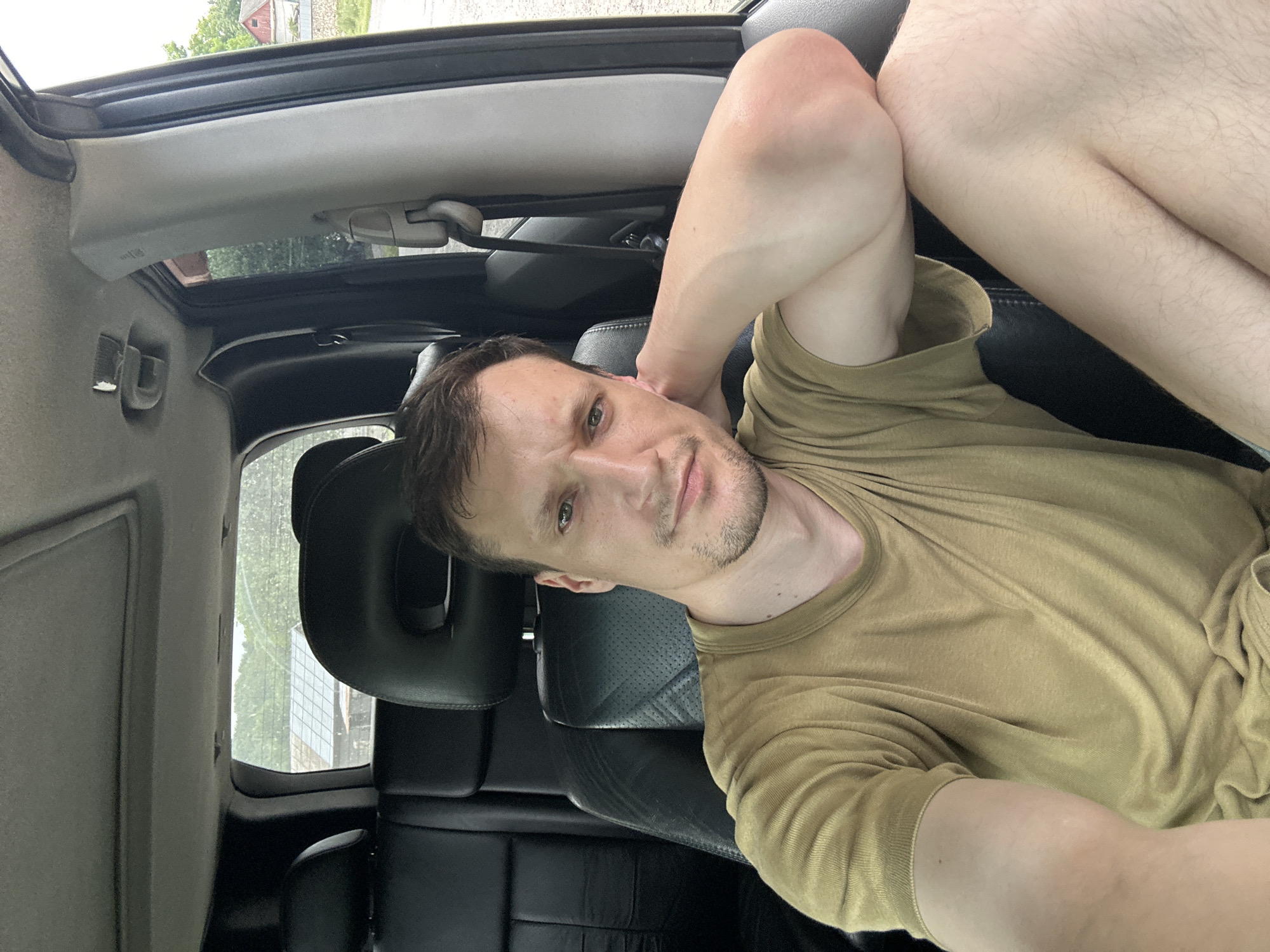 A fair-skinned adult male with short brown hair, a light stubble, and green eyes is sitting in the passenger seat of a vehicle. He is wearing a light brown t-shirt and is resting his right arm on the back of his neck, with his elbow extended. The interior of the vehicle appears to be black leather, with a sunroof visible above. Through the rear window, a small, rustic building with a tiled roof and trees are visible in the background. The scene appears to be outdoors, possibly in a rural area. The location is identified as Makiv, Ukraine.