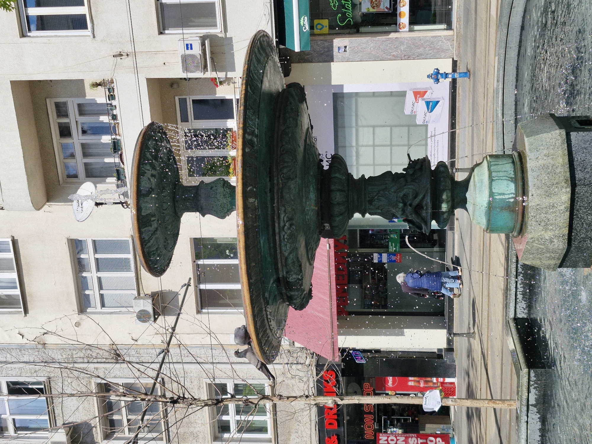 Here is a description of the image:
The image shows a fountain in Sofia, Bulgaria. The fountain has two large, circular tiers, with water cascading down. On the ground is a water-filled stone basin. A tree, likely barren, is in the foreground. In the background are several storefronts and apartments. One of the stores appears to be called "Salateria", while another is a "Non Stop" shop. A pedestrian, wearing a blue jacket and jeans, is standing on the sidewalk near the fountain. The photo appears to be taken on a sunny day. The image is rotated for some reason.