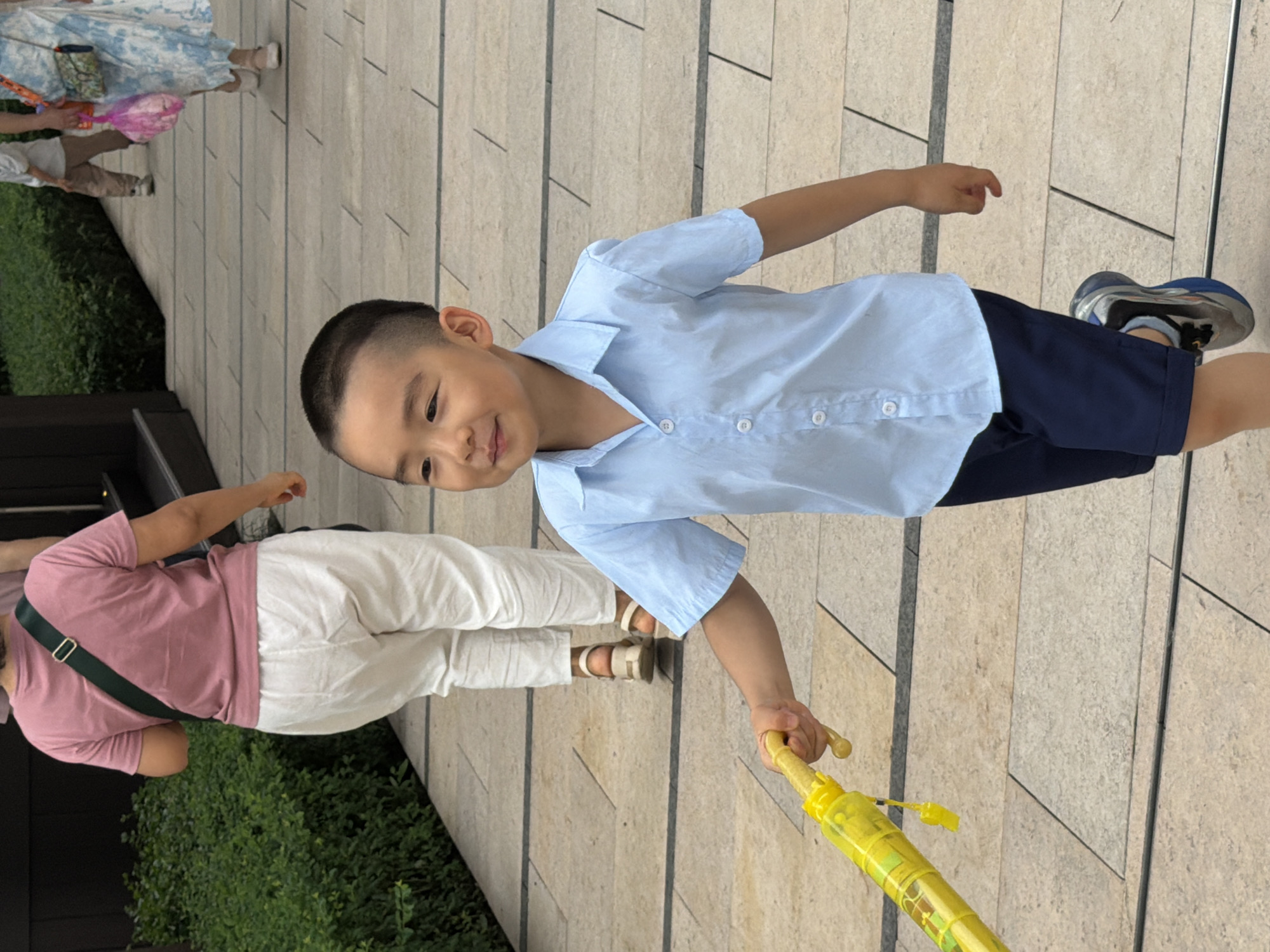 In Yuzhong District, China, a young East Asian boy with a short haircut is walking on a paved pathway outdoors. He is wearing a light blue short-sleeved collared shirt and dark blue shorts. He is holding a bright yellow umbrella with a cartoon print. The boy is looking to his left with a slight smile and his right arm is extended forward, gripping the umbrella handle. His left arm is bent at the elbow, with his hand slightly open. His legs are in motion, suggesting he is walking or running. In the background, to the left, a person wearing a pink t-shirt and white pants is walking away from the camera. Further in the background, other individuals are walking, including a person in a blue and white patterned dress carrying a pink bag. The pathway is made of rectangular light-colored stone tiles, with dark lines separating them. Green foliage is visible on the left side of the pathway.
