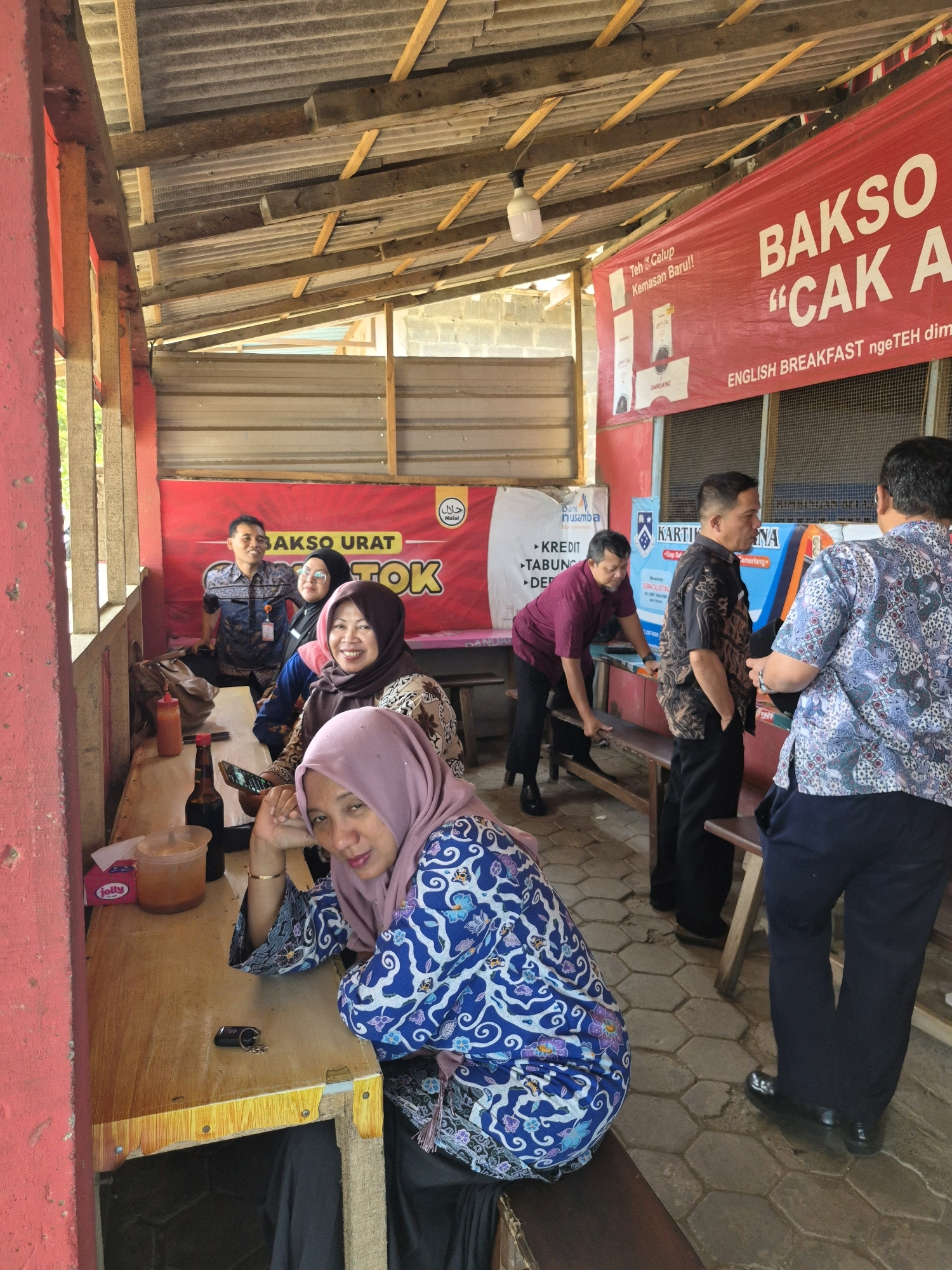 The image depicts an outdoor seating area of a food stall, likely in Purwodadi, Indonesia, characterized by a rustic wooden roof with corrugated tin sheeting supported by wooden beams. A large red banner with white text reading "BAKSO CAK A" is prominently displayed, along with other Indonesian text and logos, including a "Halal" symbol.
Several individuals are present, mostly seated at a wooden table. In the foreground, a woman wearing a light purple hijab and a blue and white patterned top is seated and looking towards the camera, with her hand resting on her chin. To her right, another woman in a darker hijab and a patterned top is also seated, smiling. Behind them, a man in a patterned blue shirt and dark pants is smiling and standing. Further back, a man in a maroon shirt and dark pants is interacting with a table, and two other men in patterned shirts and dark pants are standing and engaged in conversation.
On the table in the foreground, there are several items including a bottle of soy sauce, a container of chili sauce, a packet of tissues, and a glass of iced tea. The floor is paved with hexagonal concrete blocks. The overall scene suggests a casual dining environment at a local eatery.
