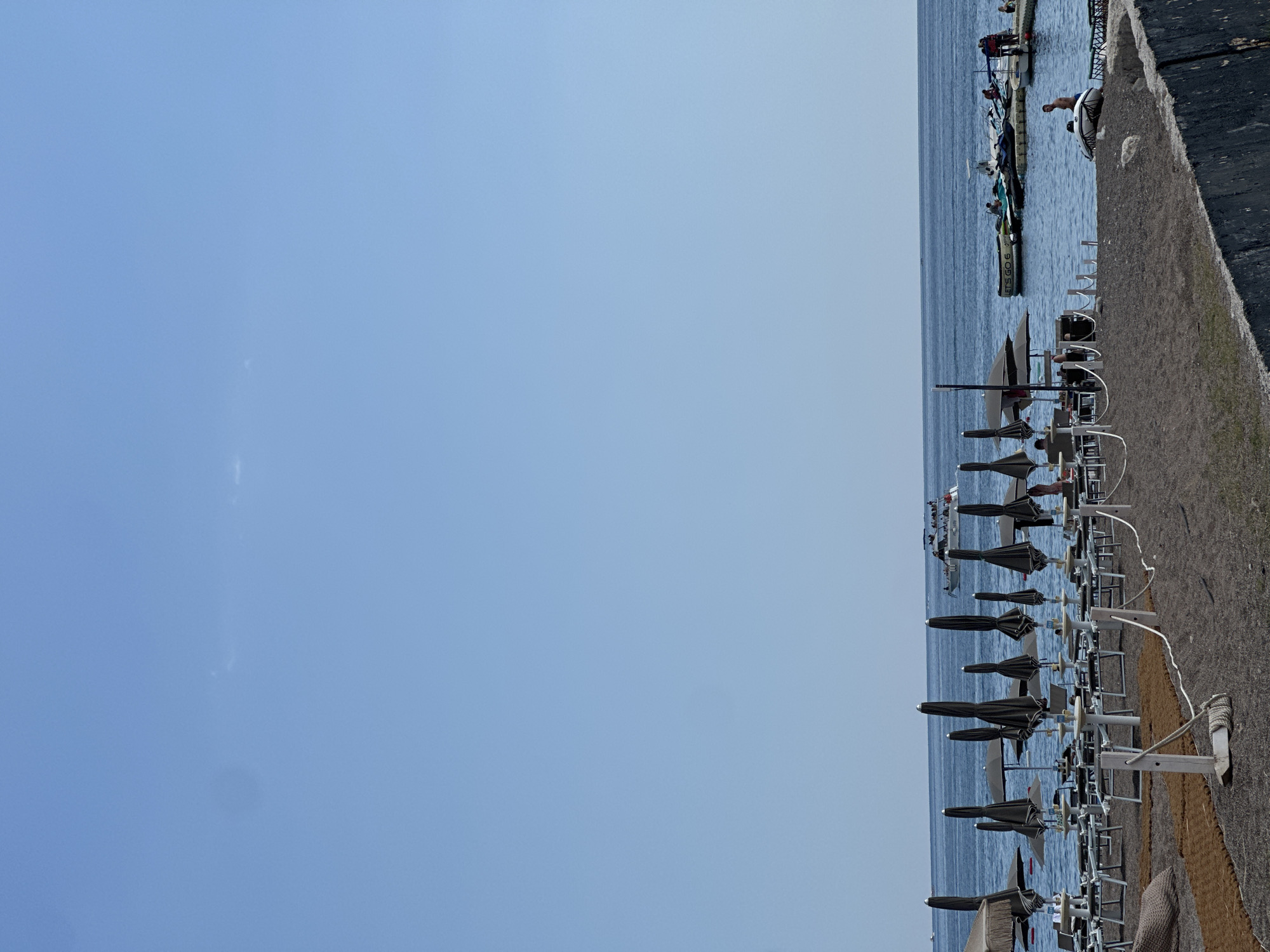 This image captures a waterfront scene in Boreti, Montenegro. In the foreground, a row of closed beach umbrellas and lounge chairs are neatly arranged on a dark, pebbly shore. Beyond the beach, the calm, blue water of the sea stretches to the horizon. A short distance away, a dock or pier extends into the water, where several small boats are moored. People can be seen on the dock and in the boats, suggesting a leisurely day at the beach or a recreational boating activity.
The sky is a clear, light blue, indicating fair weather and likely daytime, possibly late morning or early afternoon given the brightness. The overall atmosphere appears peaceful and relaxed, typical of a seaside resort. There is no discernible text visible in the image. The composition, with the vast expanse of sky on the left and the organized beach setup and water on the right, creates a sense of spaciousness and tranquility.