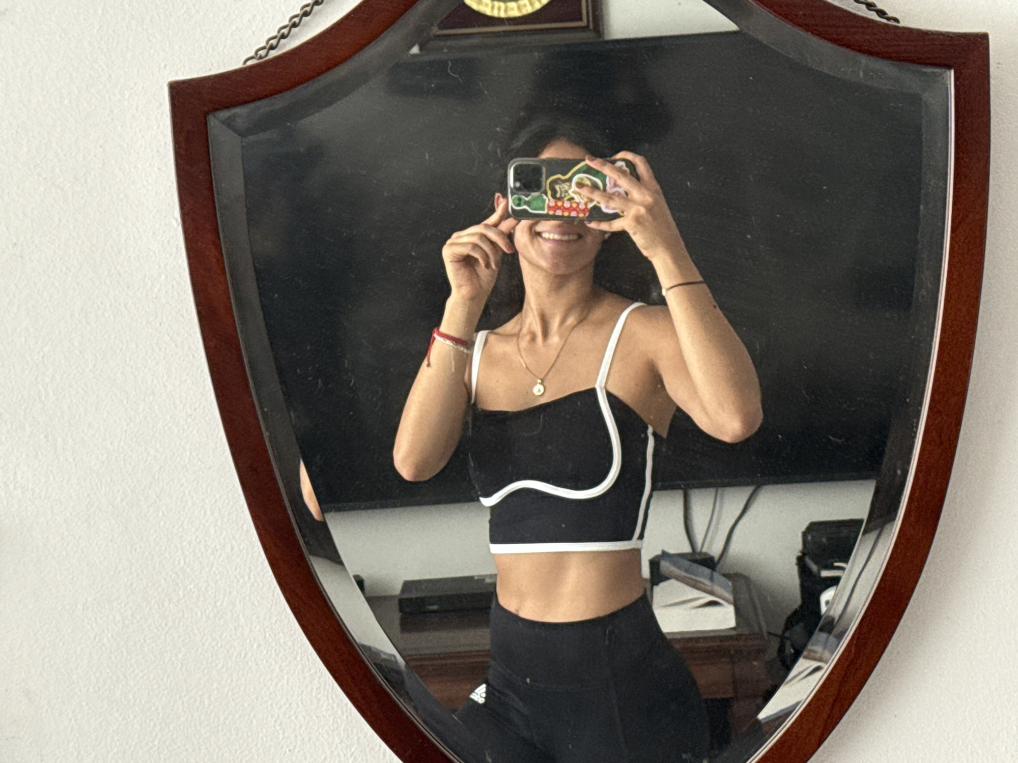 A woman is taking a picture of herself in a shield-shaped mirror. She is wearing a black and white sports bra, black leggings, a gold necklace, and a red bracelet. The mirror is hanging on a white wall. Behind the woman in the mirror's reflection, there is a table with electronic devices on it and a framed image on the wall. There is no evidence in the image to suggest the location is in the city of Puebla, Mexico.