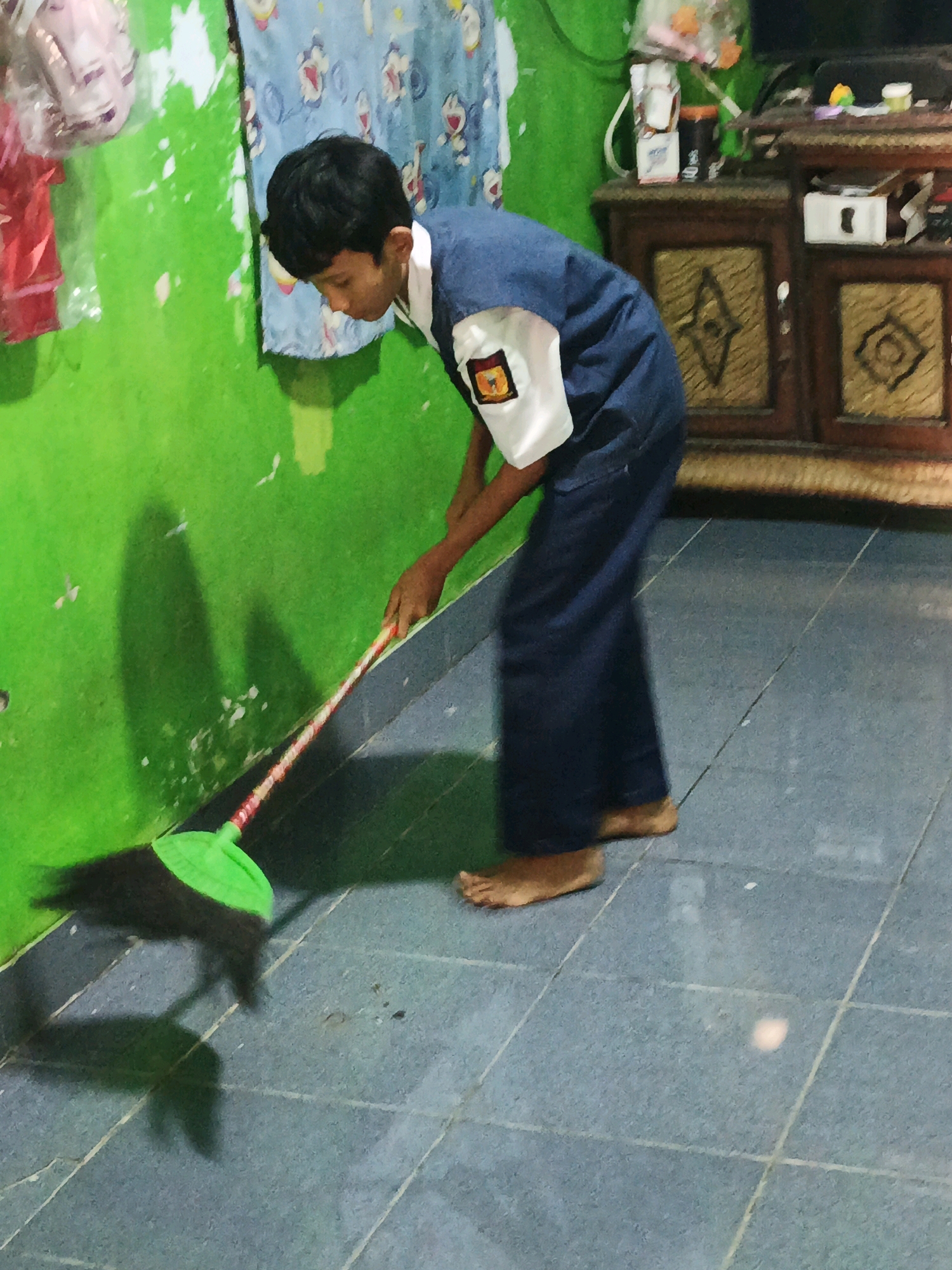 A young boy, likely a student given his uniform, is sweeping a floor with a green broom. He is barefoot and wearing dark blue pants and a blue shirt with a white collar and a patch on the left sleeve. The floor is tiled in a light blue color. To the left of the boy is a bright green wall, which appears to be textured or peeling in places. There are some red and pink items hanging on the wall to the far left. Behind the boy, to the right, is a wooden cabinet with decorative panels. Above the cabinet, there are some objects on a shelf, including what looks like a television. The overall impression is of an indoor residential setting, possibly in Santing, Indonesia, where cleaning is being done. The lighting suggests an indoor environment, with shadows cast on the floor and wall.