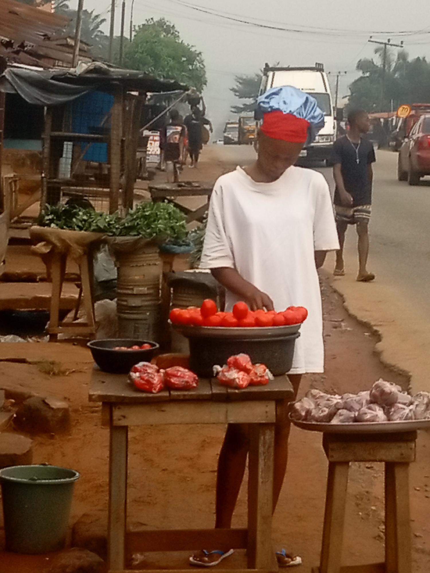 A young woman is standing by a table full of tomatoes, likely in Nkpor, Nigeria. She is wearing a white T-shirt and a red and blue head covering. She is looking down at the tomatoes in a black bucket. On the table, there is also a black bowl of tomatoes and some other packaged items. There is a tray of wrapped meat on a small stool to the right. Behind her, a young man is walking on the sidewalk, and vehicles are visible on the road. There is also a simple wooden structure to the left with green vegetables on top. The ground is reddish-brown, and the weather appears overcast.