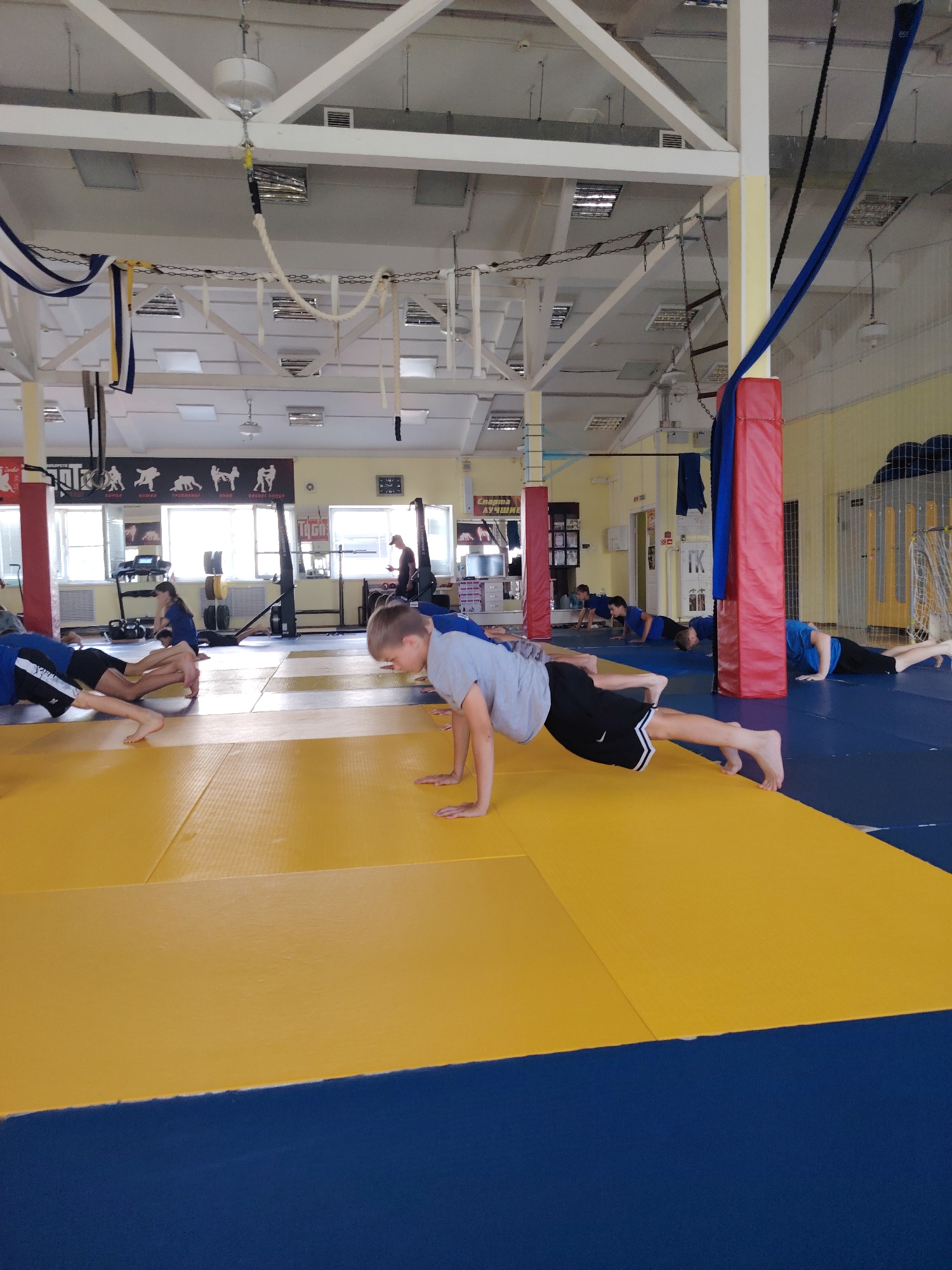 In a gymnasium in Chaikovsky, Russia, several individuals are performing push-ups on yellow mats. The room is spacious with a high ceiling and exposed white beams. Various gymnastic equipment, including ropes and a net, are suspended from the ceiling. Posters depicting wrestling or martial arts figures are visible on the walls. In the background, windows allow natural light to enter, illuminating the scene. The individuals, primarily youths and young adults, are dressed in athletic wear, with some wearing blue tops and black shorts. The overall environment suggests a training session for a sport requiring physical exertion.