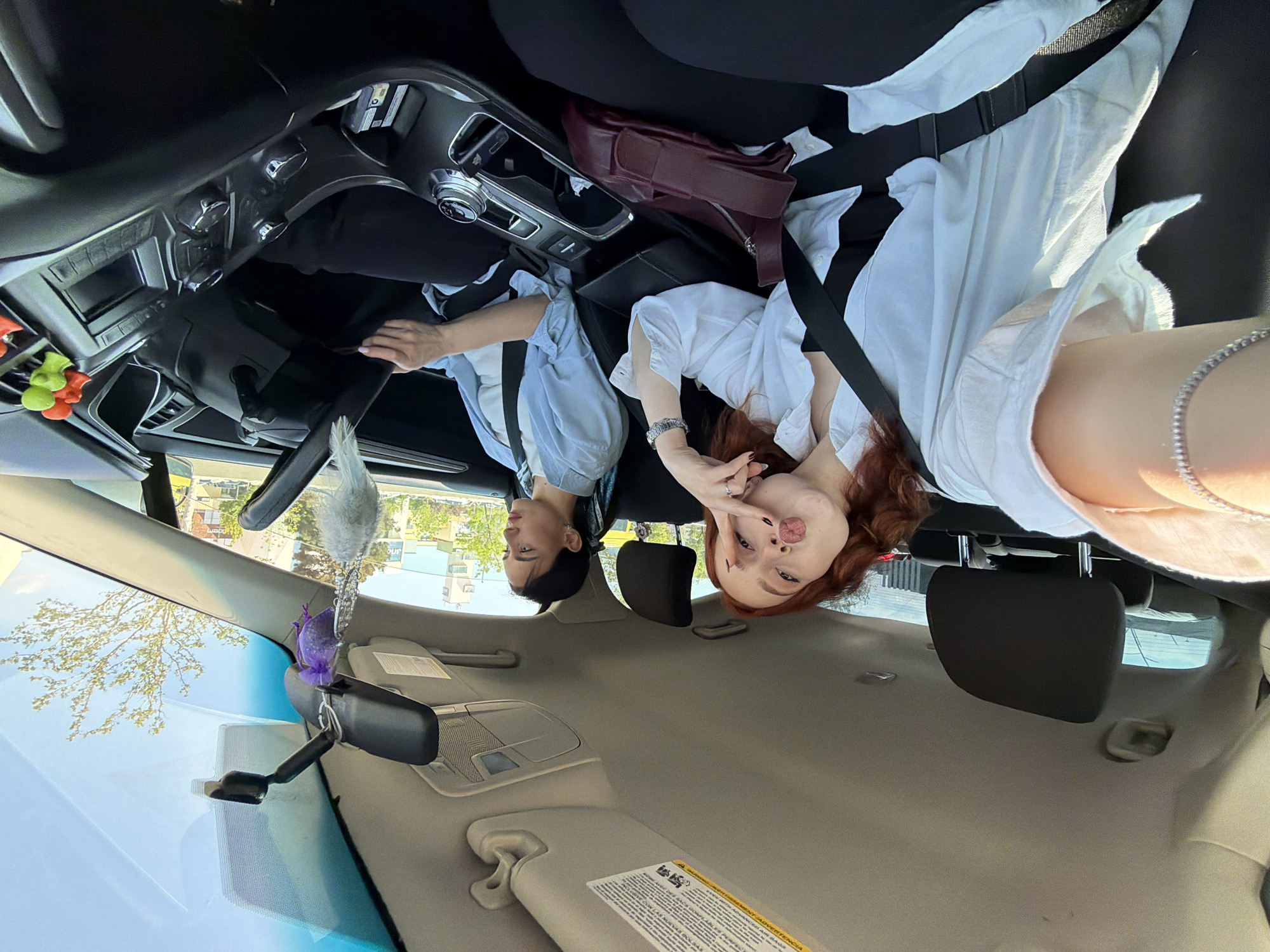 The image captures two individuals seated in the front of a car during daylight, presumably in Tbilisi, Georgia. The photo appears to be taken from the backseat, looking forward, with the subjects appearing upright after mental rotation.
On the right, a young woman with reddish-brown hair, dressed in a white collared shirt and black pants, playfully poses for the camera. She extends her tongue, puckers her lips, and forms a peace sign with her right hand. A silver bracelet is visible on her left wrist. On the left, another person with dark hair, wearing a light blue collared shirt and black pants, looks straight ahead, seemingly at the road or out the front window.
Between the two individuals on the center console, a burgundy purse or bag rests. The car's interior features are visible, including parts of the dashboard, steering wheel, and a rearview mirror from which a fluffy white ornament and a small purple fabric sachet hang. Near the dashboard on the driver's side, colorful green and yellow keychains or small figures are attached. Through the car's windows, bright daylight illuminates the scene, revealing green trees and buildings under a clear sky, indicative of sunny weather. A warning label on the passenger's sun visor is visible, showing text, including the word "ADVERTENCIA," suggesting a multi-language safety notice, likely English and Spanish. The overall atmosphere conveyed is lighthearted and relaxed.