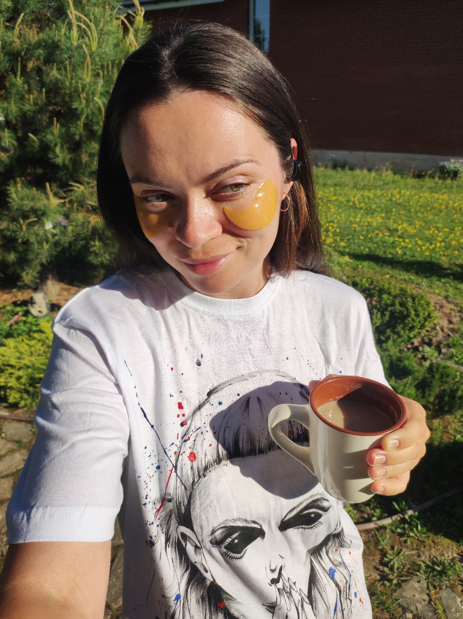 A selfie shows a light-skinned woman with dark brown hair, wearing orange eye mask patches, holding a mug of a beige liquid, likely coffee, against a backdrop of greenery. The woman wears a white t-shirt featuring a graphic print of a face and colored paint splatters. The background includes a green coniferous tree, yellow dandelions, and a brick building, likely taken in a garden or backyard. The location, Фоки, Russia is not verifiable from the image itself.