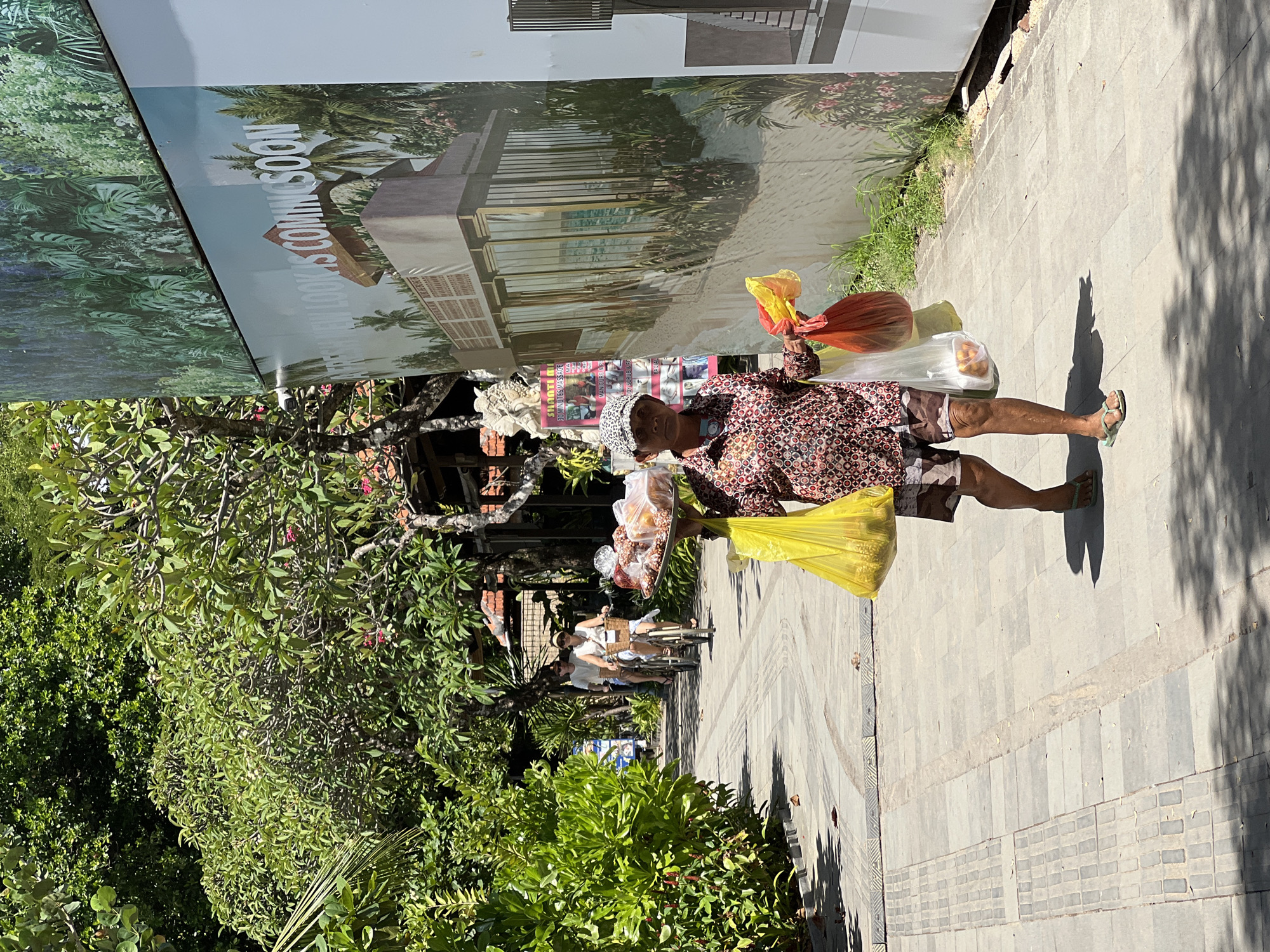 A woman is depicted walking on a paved sidewalk under bright daylight in Denpasar, Indonesia. She is the central subject, dressed in a long-sleeved patterned shirt, camouflage-print shorts, and light green flip-flops. She carries multiple plastic bags: a bright yellow one in her left hand, and a red and a clear one (containing orange-colored items, possibly fruit) in her right hand. Additionally, she appears to be balancing a flat tray with several baked goods or snacks on her head.
The scene is set in a sunny, tropical environment. To the left of the sidewalk, lush green foliage, bushes, and trees dominate the landscape. To the right, a large construction hoarding or banner features an architectural rendering of a modern building or resort surrounded by palm trees, bearing the visible text "NEW LOOK IS COMING SOON". Further behind the woman, a smaller poster or sign advertises services, with partial text readable as "SHOOT ME UP 90 MINUTES MASSAGE" and "THAI BALI".
The strong sunlight casts distinct shadows on the sidewalk, indicating a clear, bright day. In the background, two individuals are visible riding bicycles along the path. The sidewalk is constructed from light-colored pavers, with some sections featuring decorative patterns. The image captures a lively moment on a tropical street.