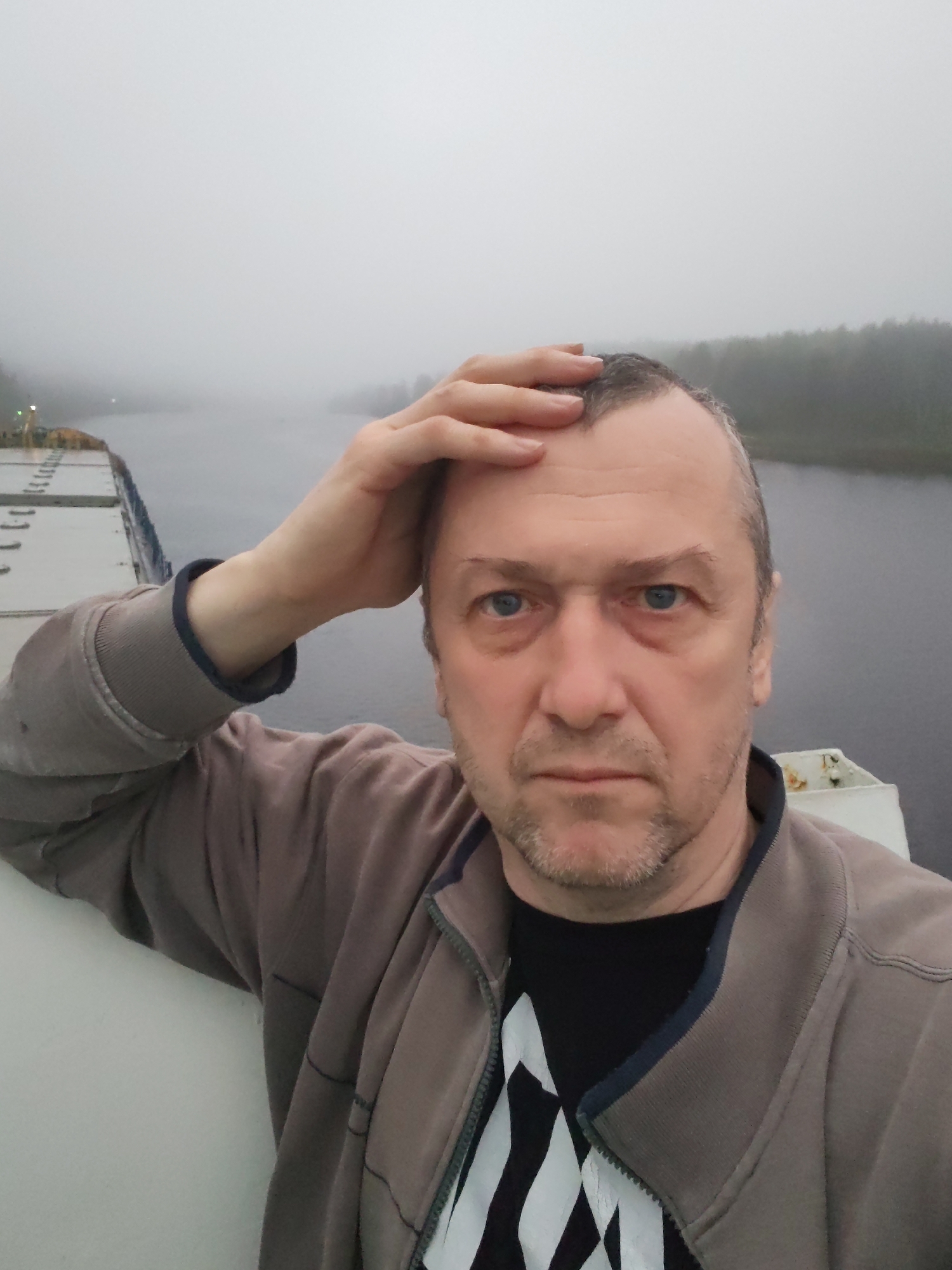 A middle-aged man is taking a selfie on a bridge or vessel. He is fair-skinned, with thinning gray hair, blue eyes, and a trimmed beard. He's wearing a gray zip-up jacket over a black shirt with a white design. His right hand is placed on his forehead.
The background is obscured by heavy fog. To the left, part of a large vessel is visible. Beyond that, a body of water stretches into the distance, bordered by a line of trees shrouded in fog.
Based on the title information, the location is likely in Volokov Most, Russia. However, this cannot be verified from the visual data in the photo. The presence of the vessel and body of water suggests the man may be on a canal or river.