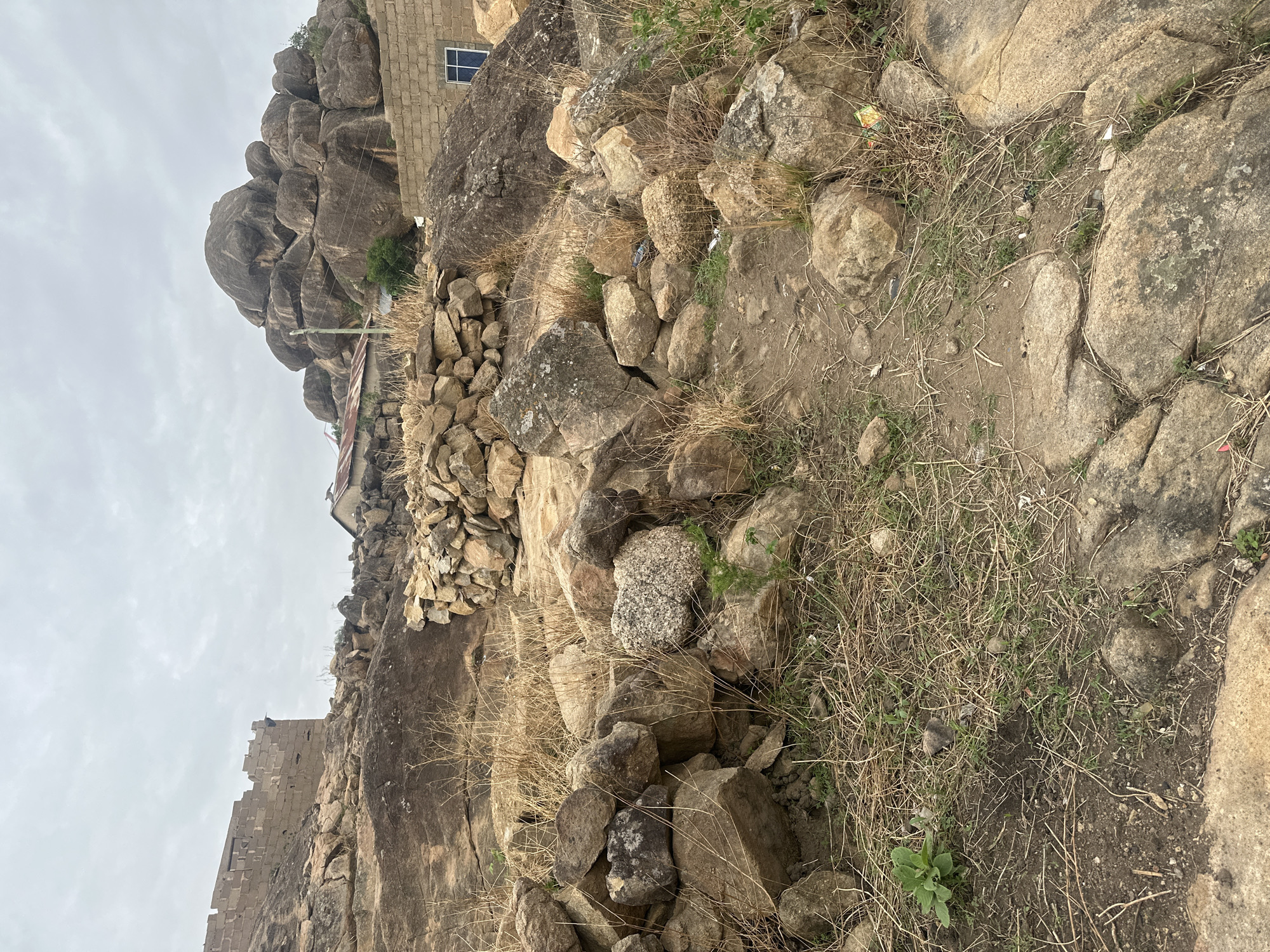 This image depicts a rocky, uneven landscape with a building constructed amongst the rocks. The foreground consists of a mix of small to medium-sized rocks, dry grass, and patches of bare earth. The building appears to be made of stone or brick, with a visible window. In the distance, there is a large, rounded rock formation. The sky is overcast. Based on the reference to Jos, Nigeria, the scene is likely located in an area characterized by rocky hills and scattered vegetation, where buildings are often integrated into the natural landscape.